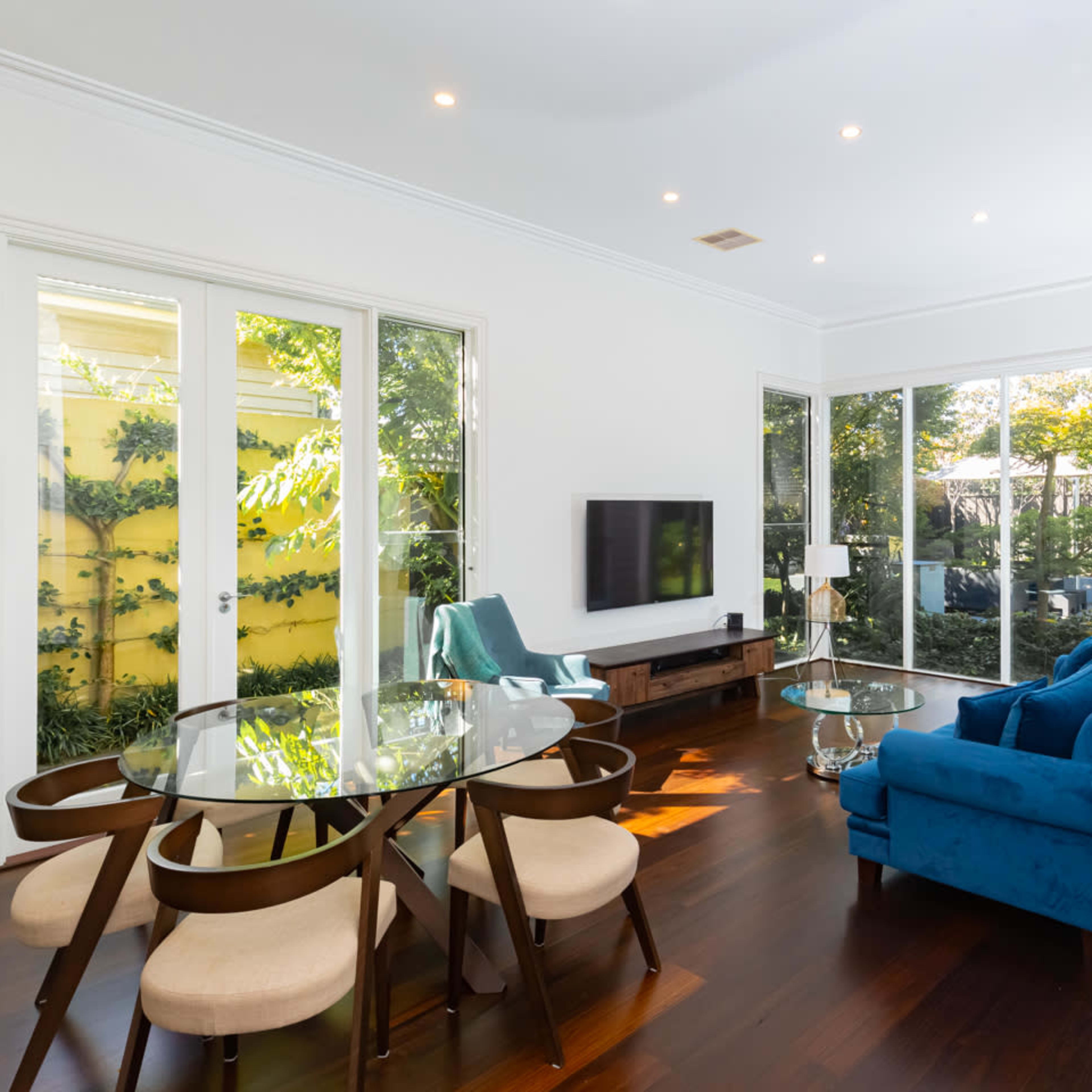 A modern living room features a glass dining table, a blue sofa, and large windows displaying a garden view.