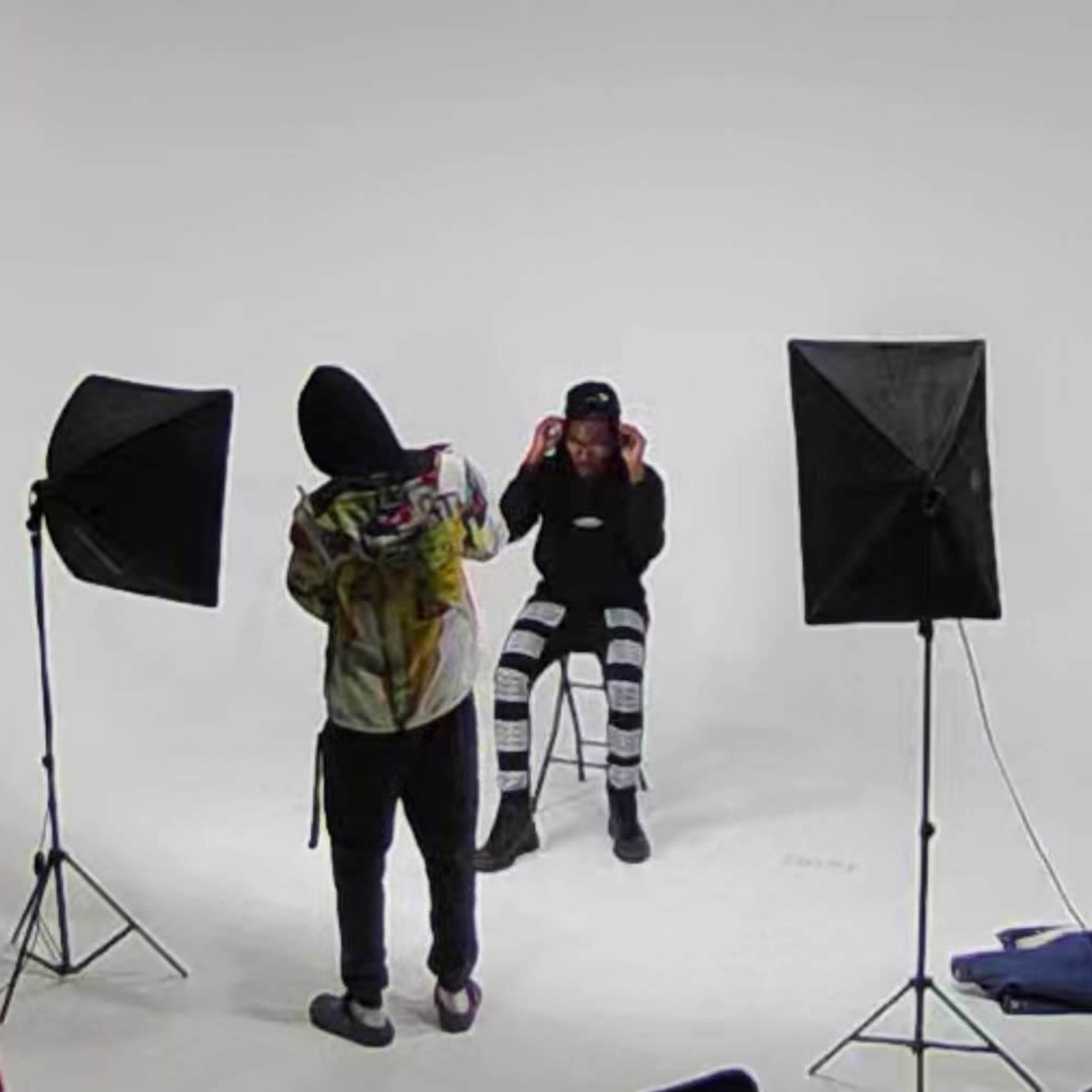 A person wearing a hoodie prepares to photograph another person seated on a stool, with studio lighting set up around them.