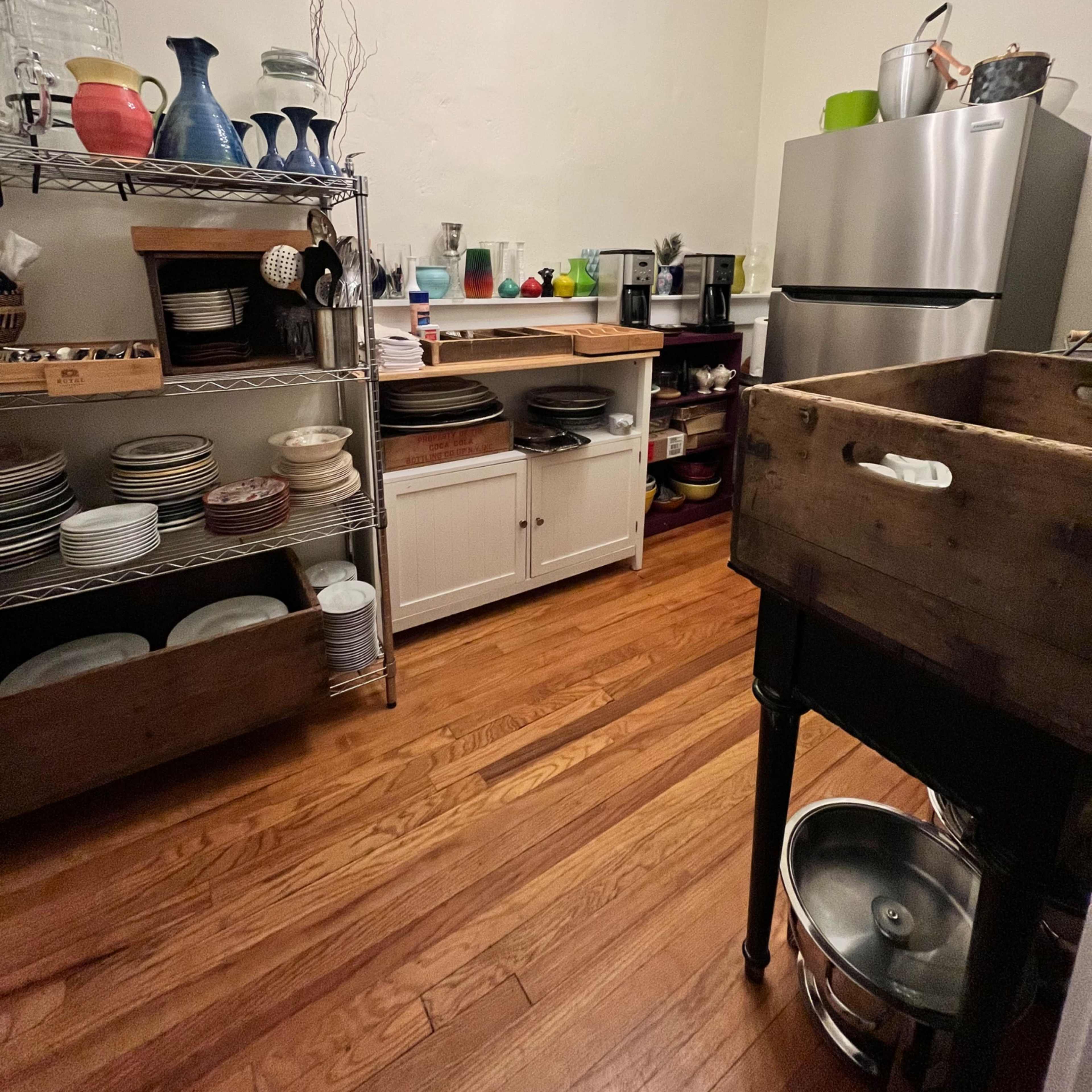 A kitchen with wooden floors, shelves filled with dishes, a counter with various kitchen appliances, and a large sink in the foreground.