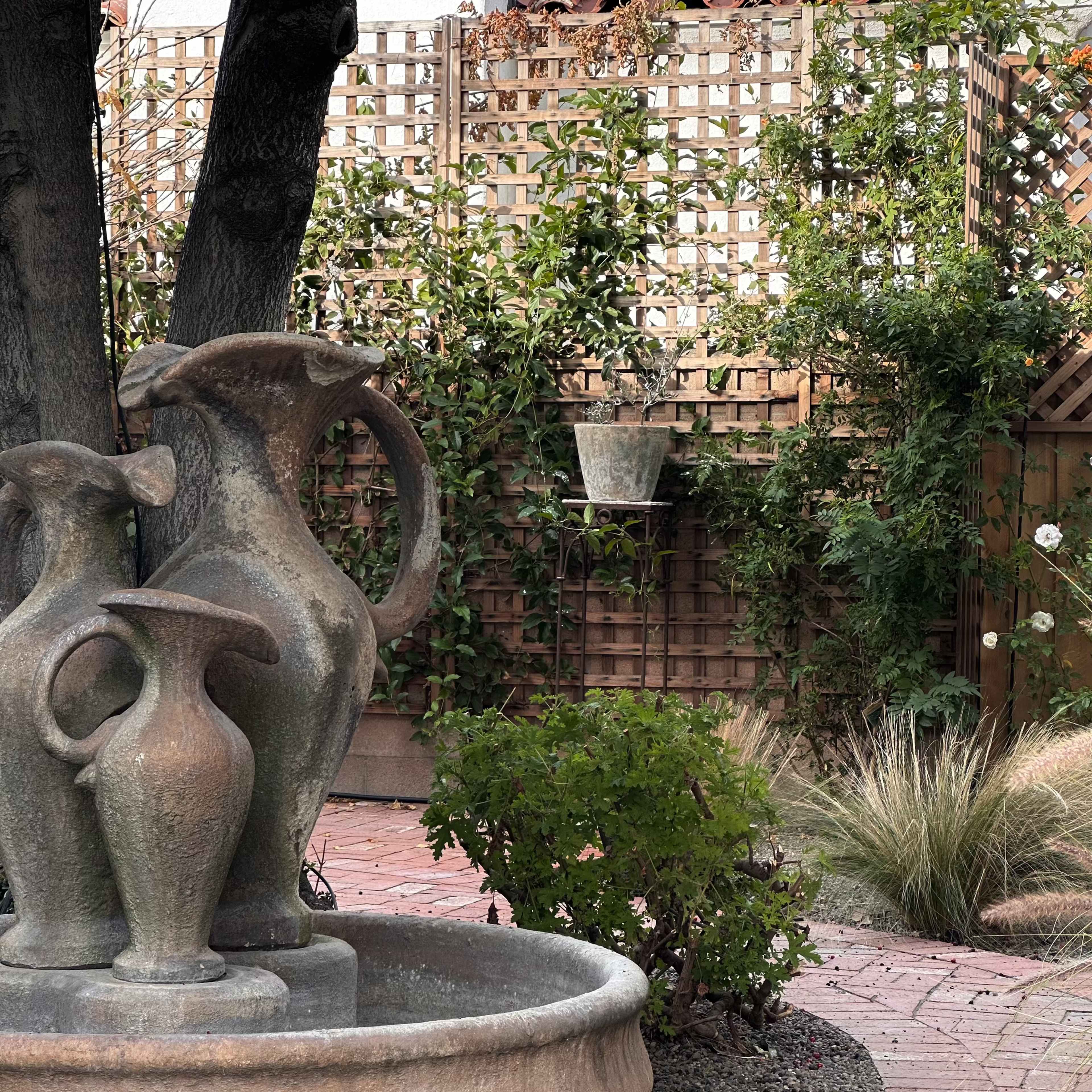 A garden scene featuring a stone fountain with several pitchers and a backdrop of a wooden lattice covered in greenery.
