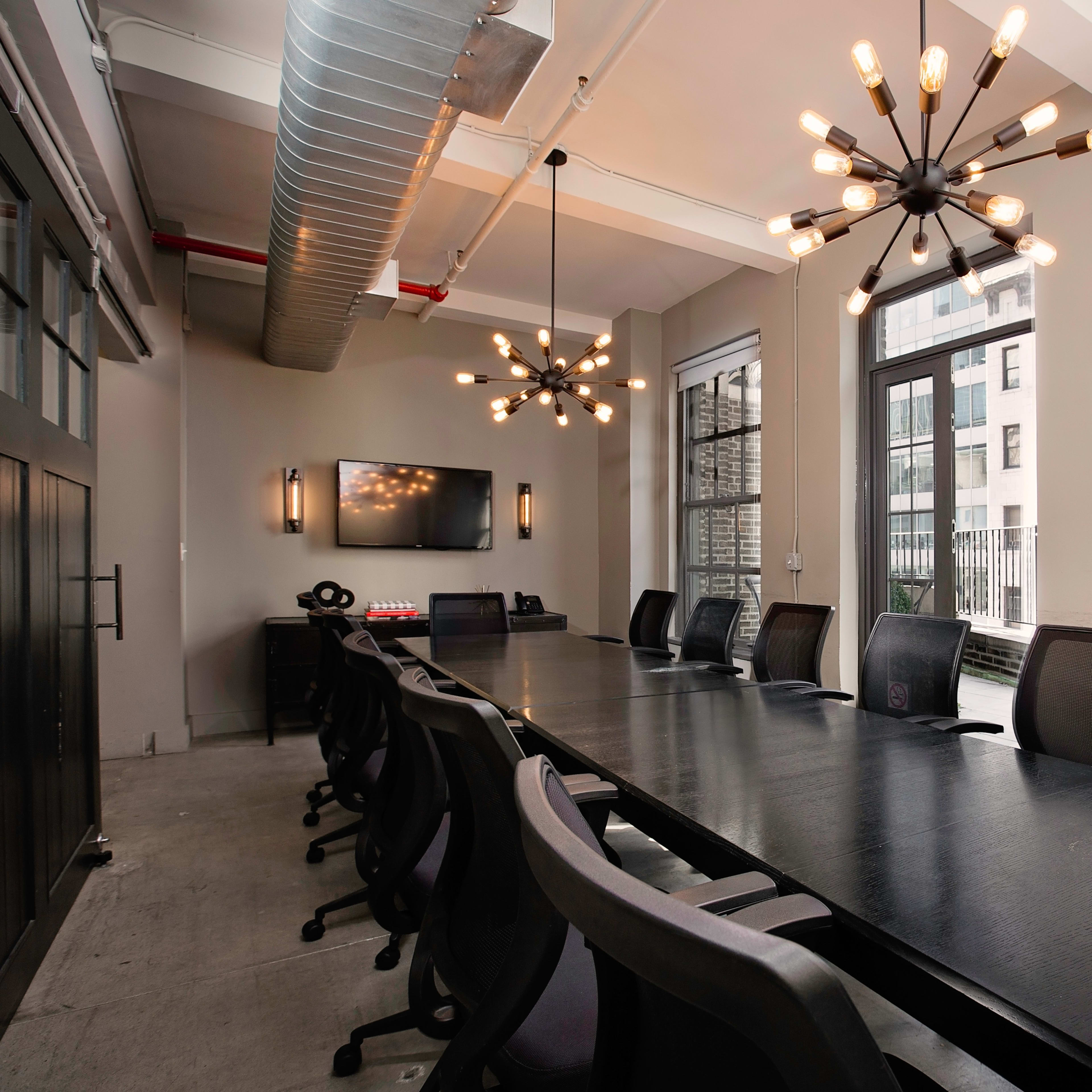 A modern conference room featuring a long black table, black ergonomic chairs, and two unique light fixtures, with large windows providing views of a cityscape.