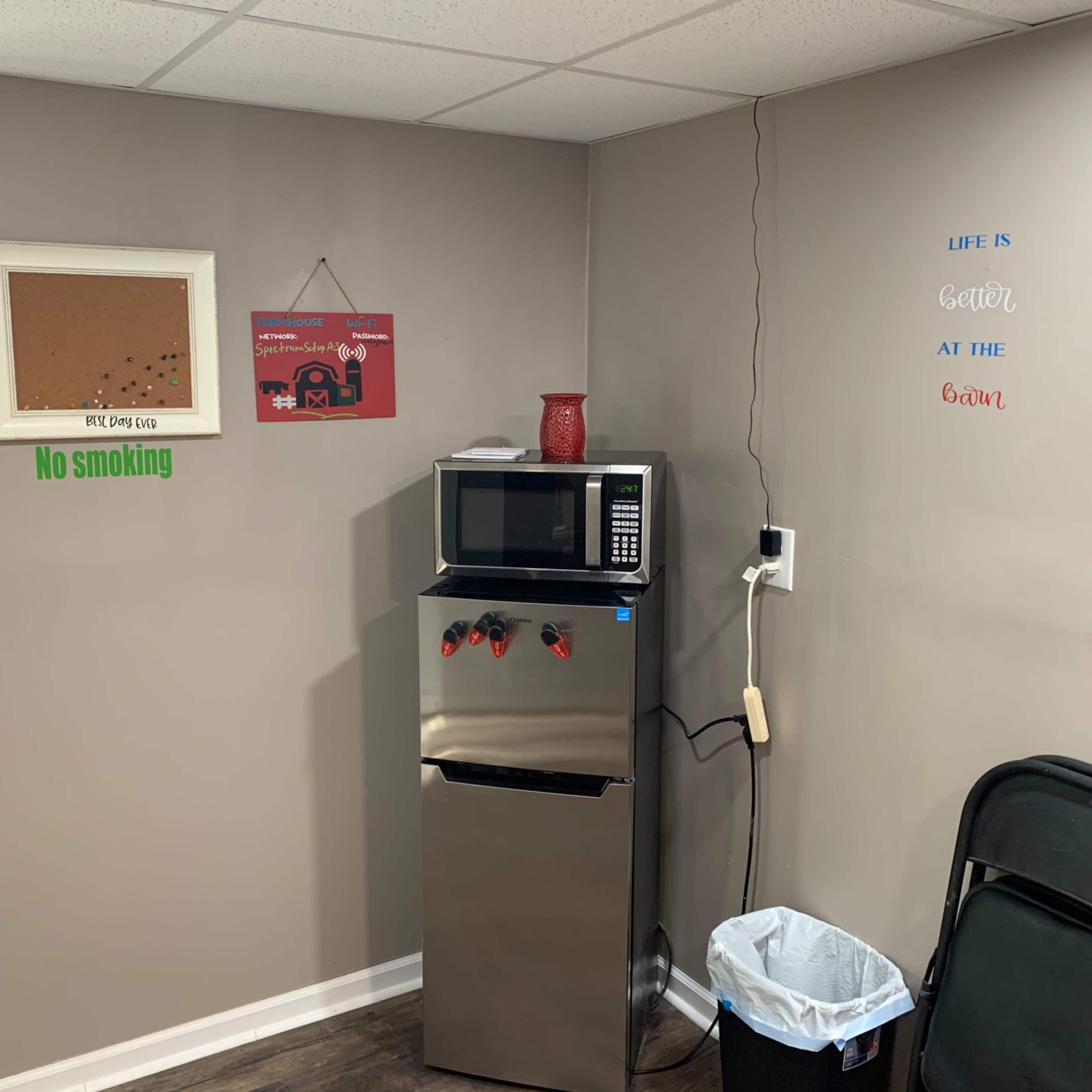 A small indoor area featuring a stainless steel refrigerator, a microwave on top, a corkboard on the wall, and a wastebasket beside a folding chair.