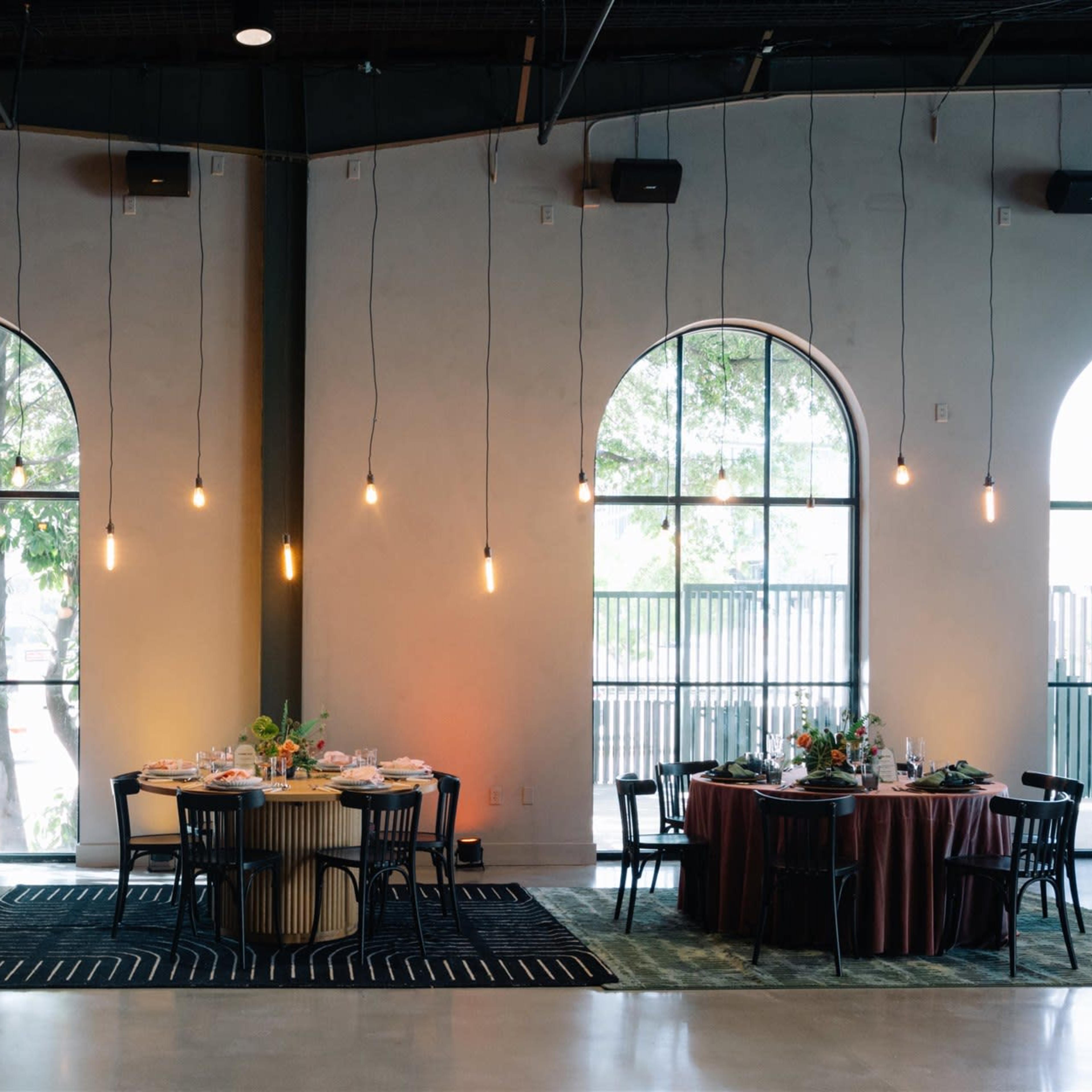 A dining area with two round tables elegantly set, surrounded by large arched windows and illuminated by hanging pendant lights.