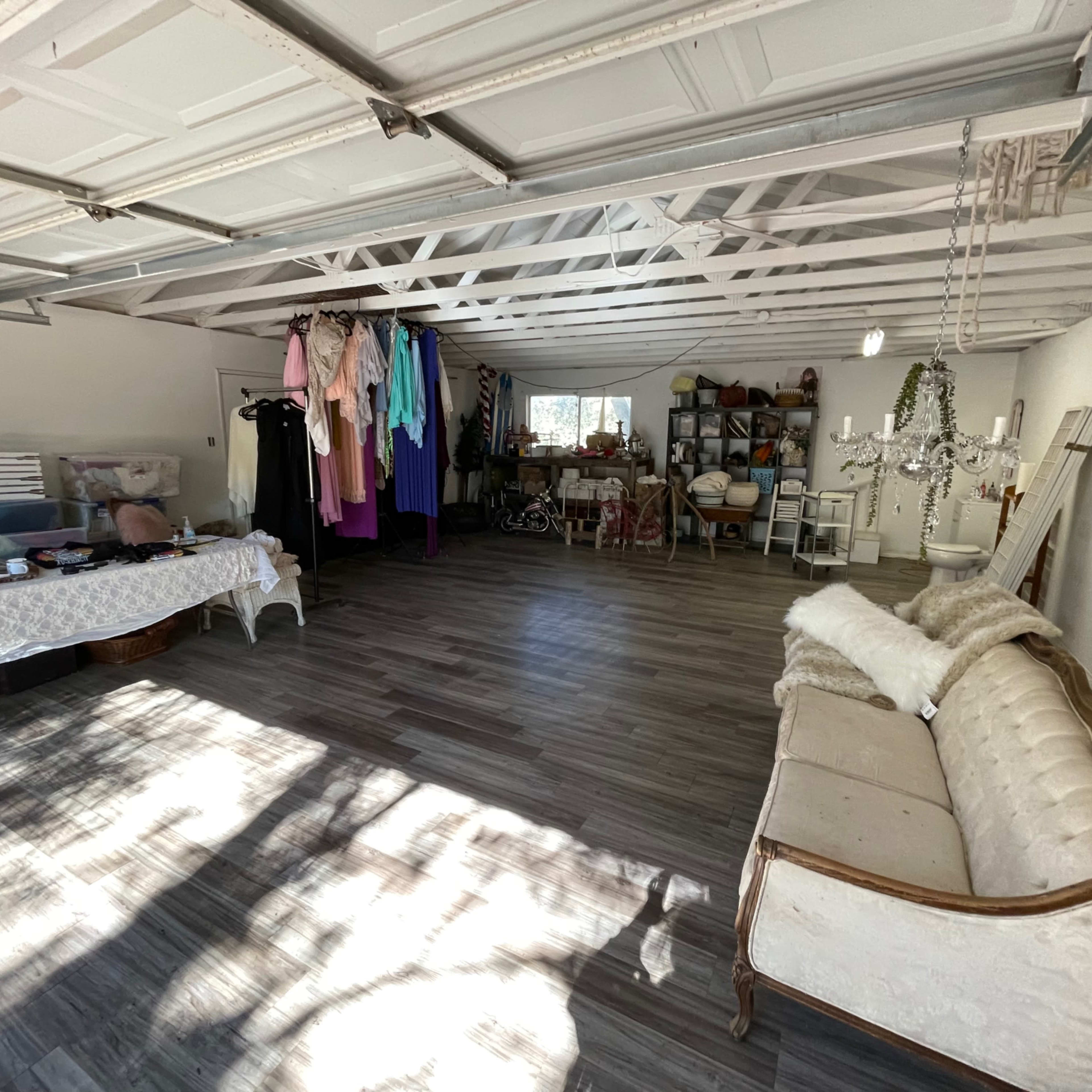 A spacious garage interior with a variety of clothing hanging on racks, a table covered with items, and a vintage couch positioned along one wall.