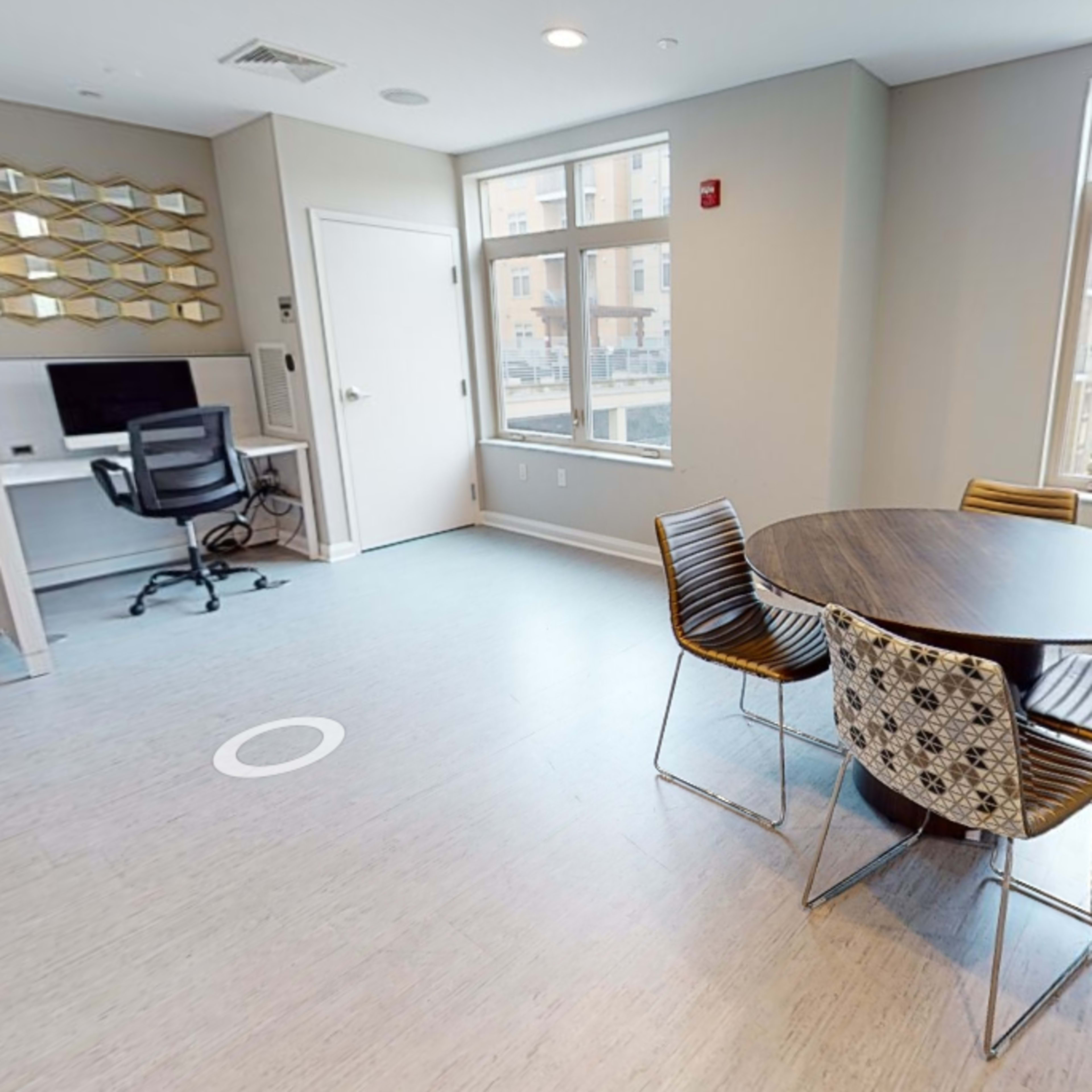 A modern room with a round table and four chairs, a desk with a chair in the corner, and large windows allowing natural light to enter.