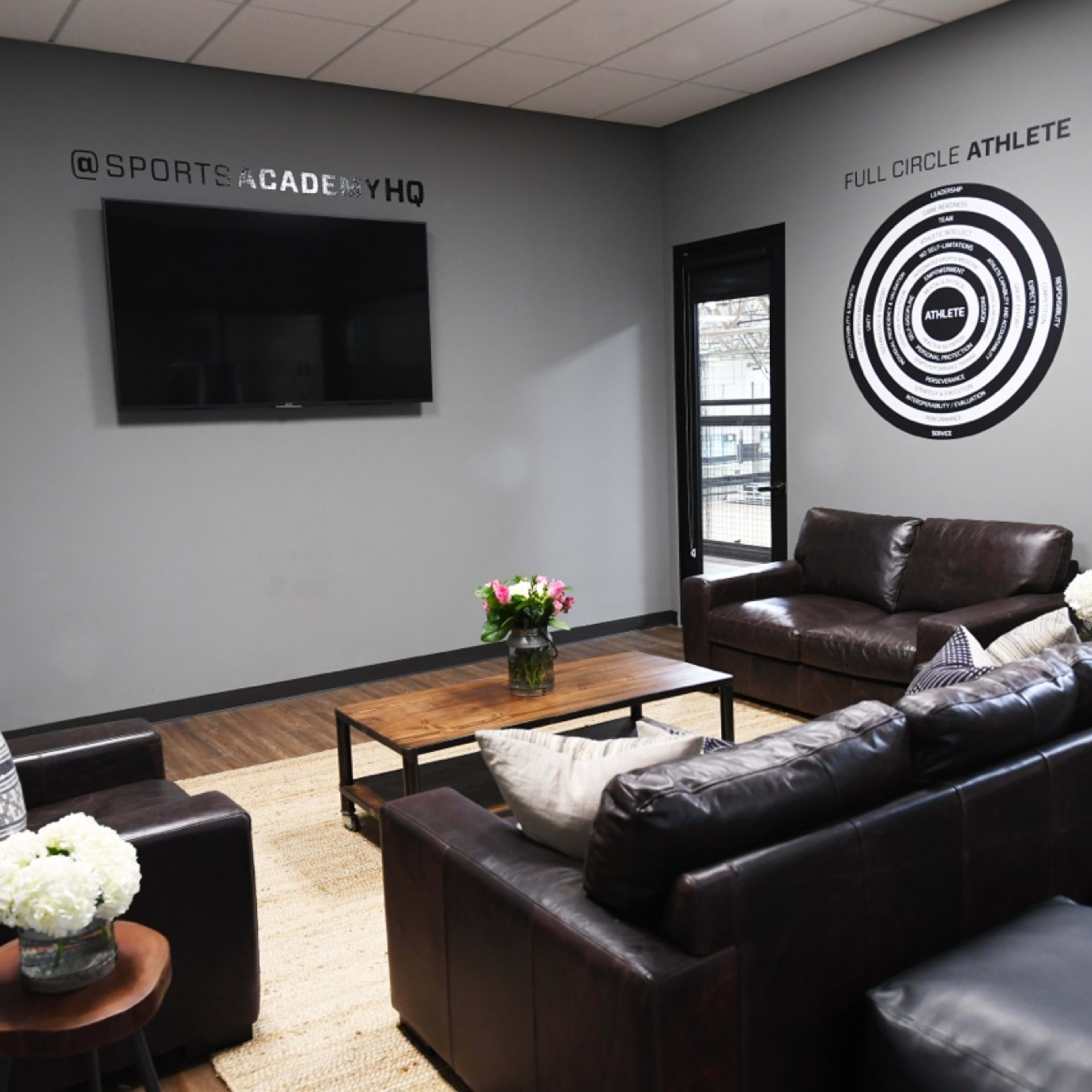 A modern lounge area with two dark leather sofas, a coffee table, a television mounted on the wall, and decorative wall art, all situated in a bright room with large windows.
