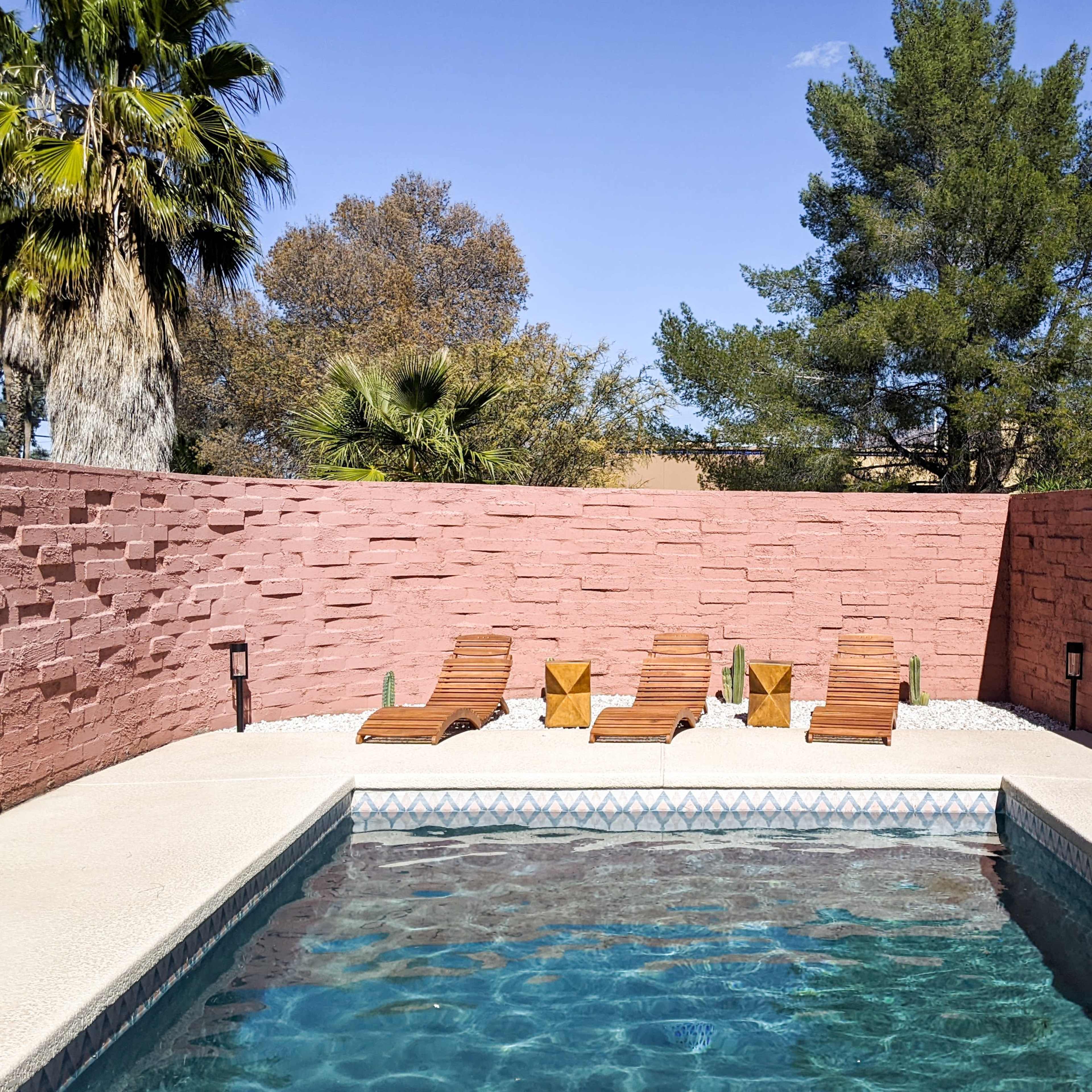 A swimming pool surrounded by a pink stone wall and four wooden lounge chairs beside it.