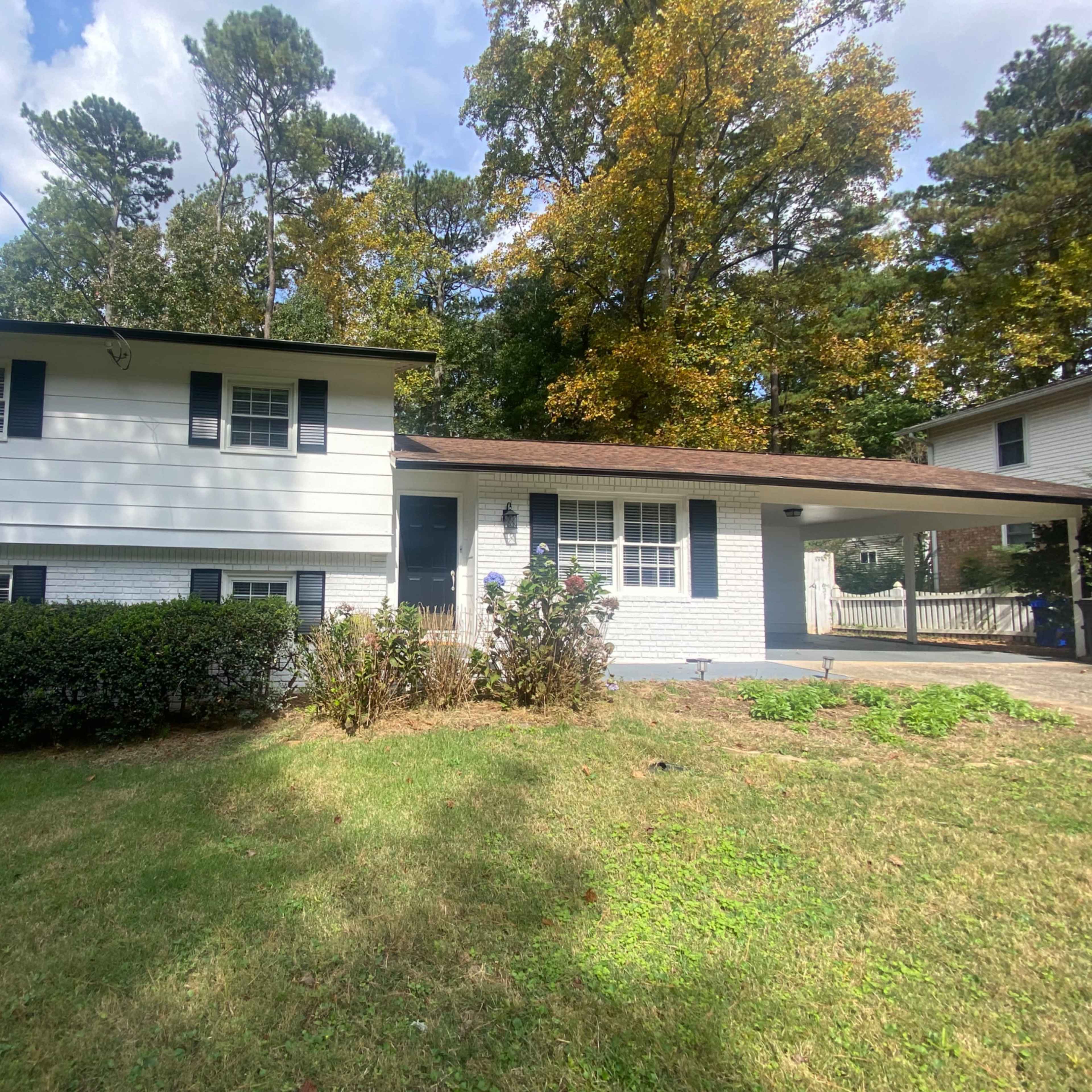 A two-story white house with a brown roof and black shutters is surrounded by green grass and trees.
