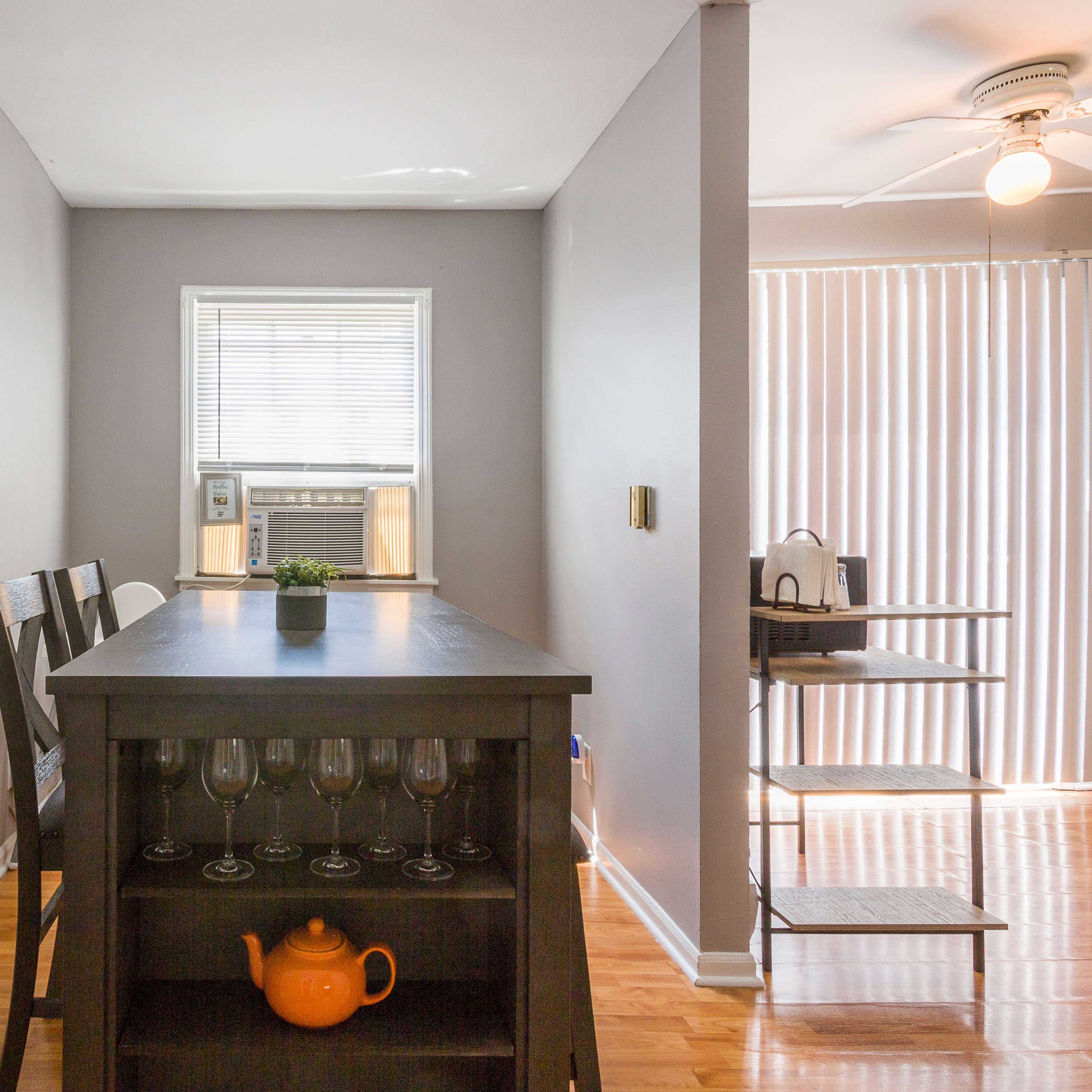 A simple dining area with a wooden table, four chairs, and a small shelf with a teapot and glasses, set against gray walls and a window with blinds.
