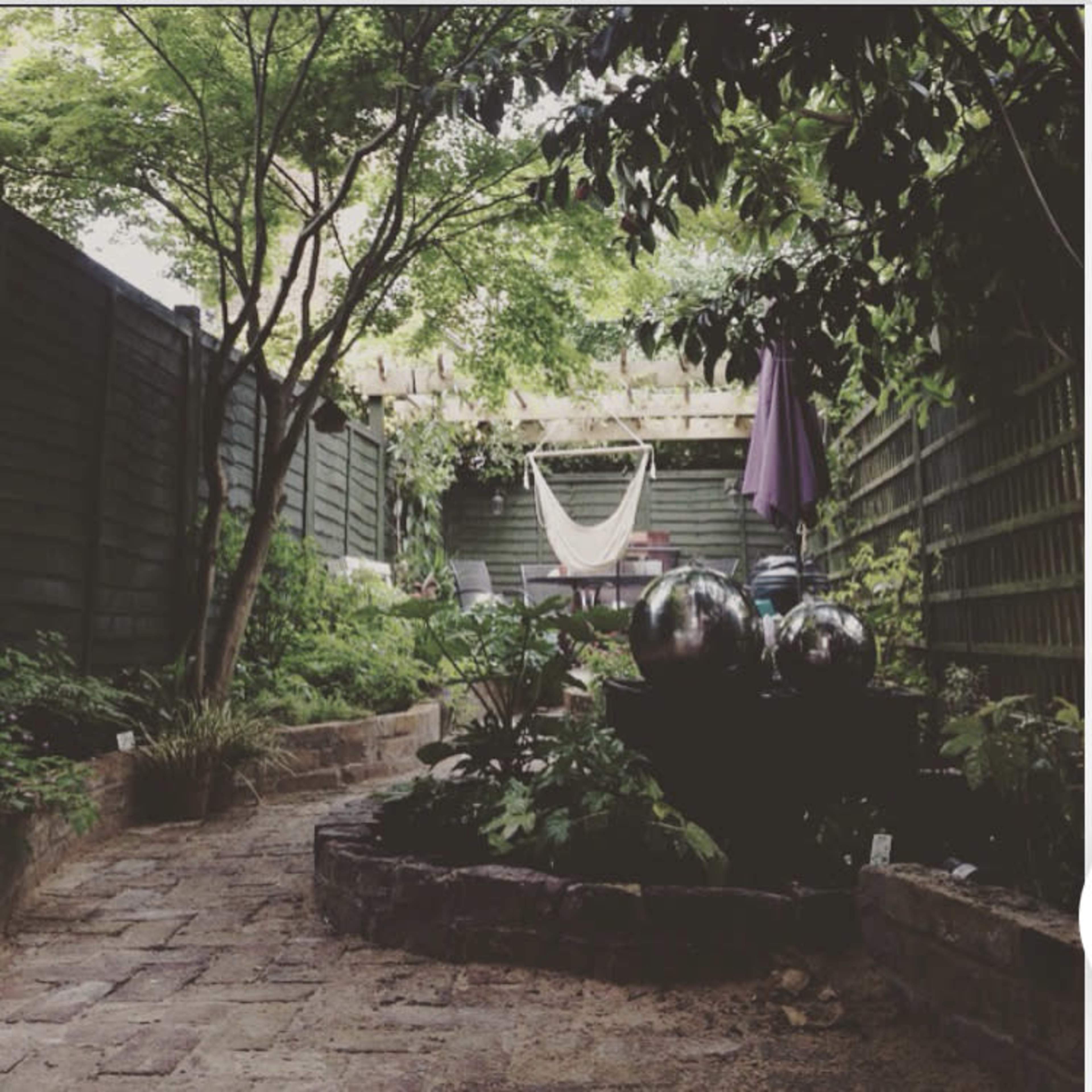 A garden pathway leading to a seating area with a hammock and decorative round fountains amidst greenery and a wooden fence.