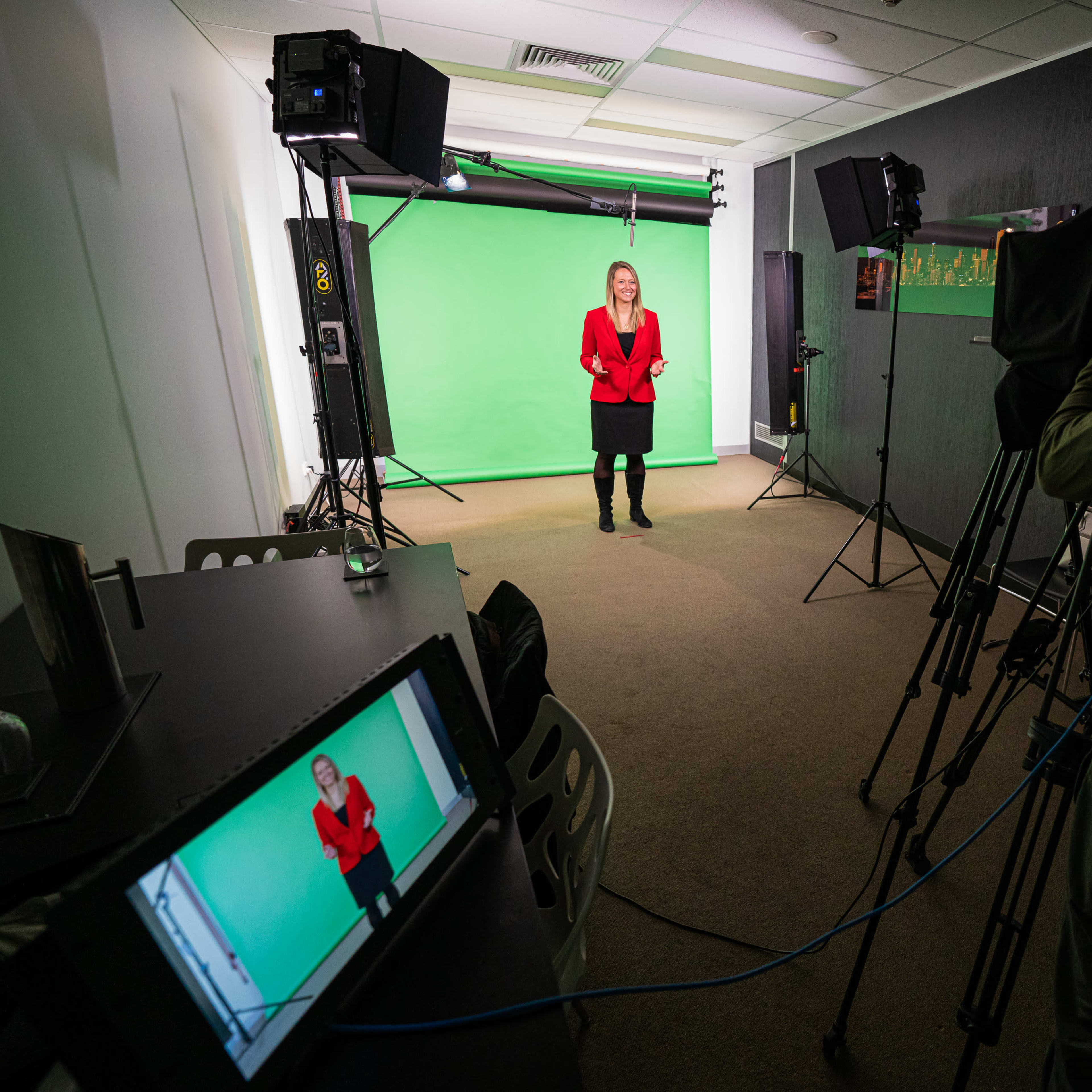 A woman in a red blazer stands in front of a green screen, with a camera and crew capturing her from the opposite side of the room.