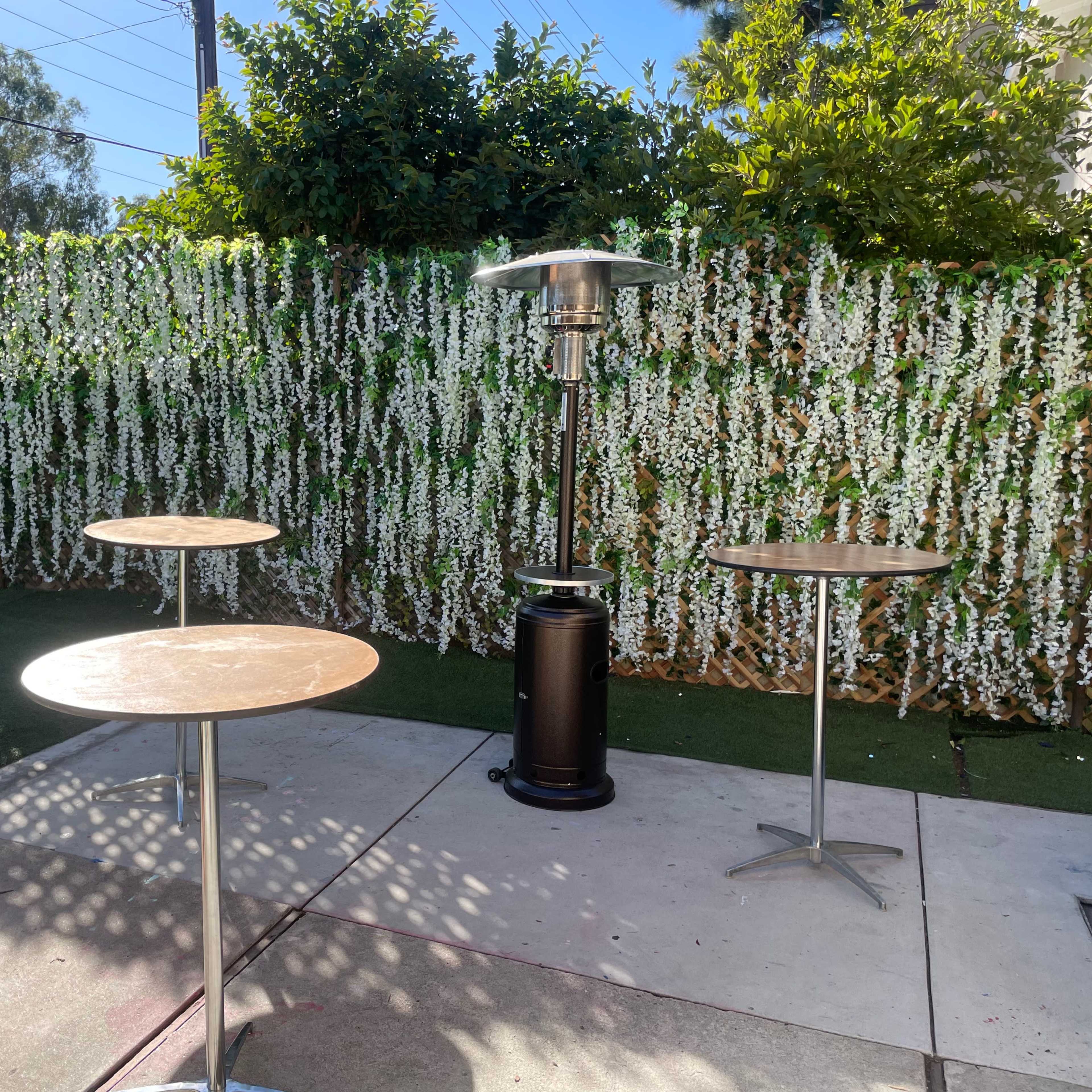 An outdoor space with two round metal tables, a patio heater, and a backdrop of lush greenery covered in white flowers.