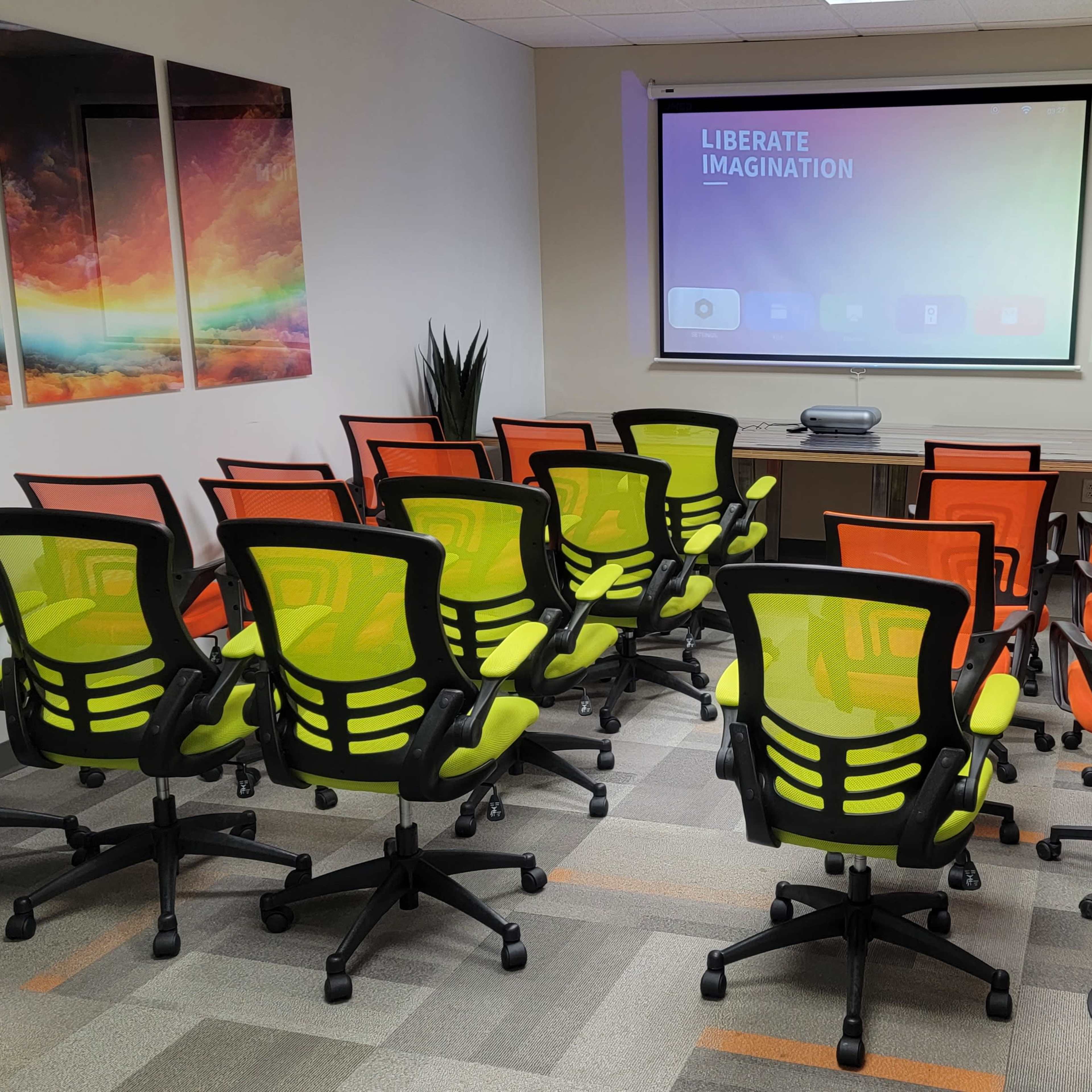 A conference room with rows of green and orange chairs facing a wall-mounted projector screen, flanked by two plants.