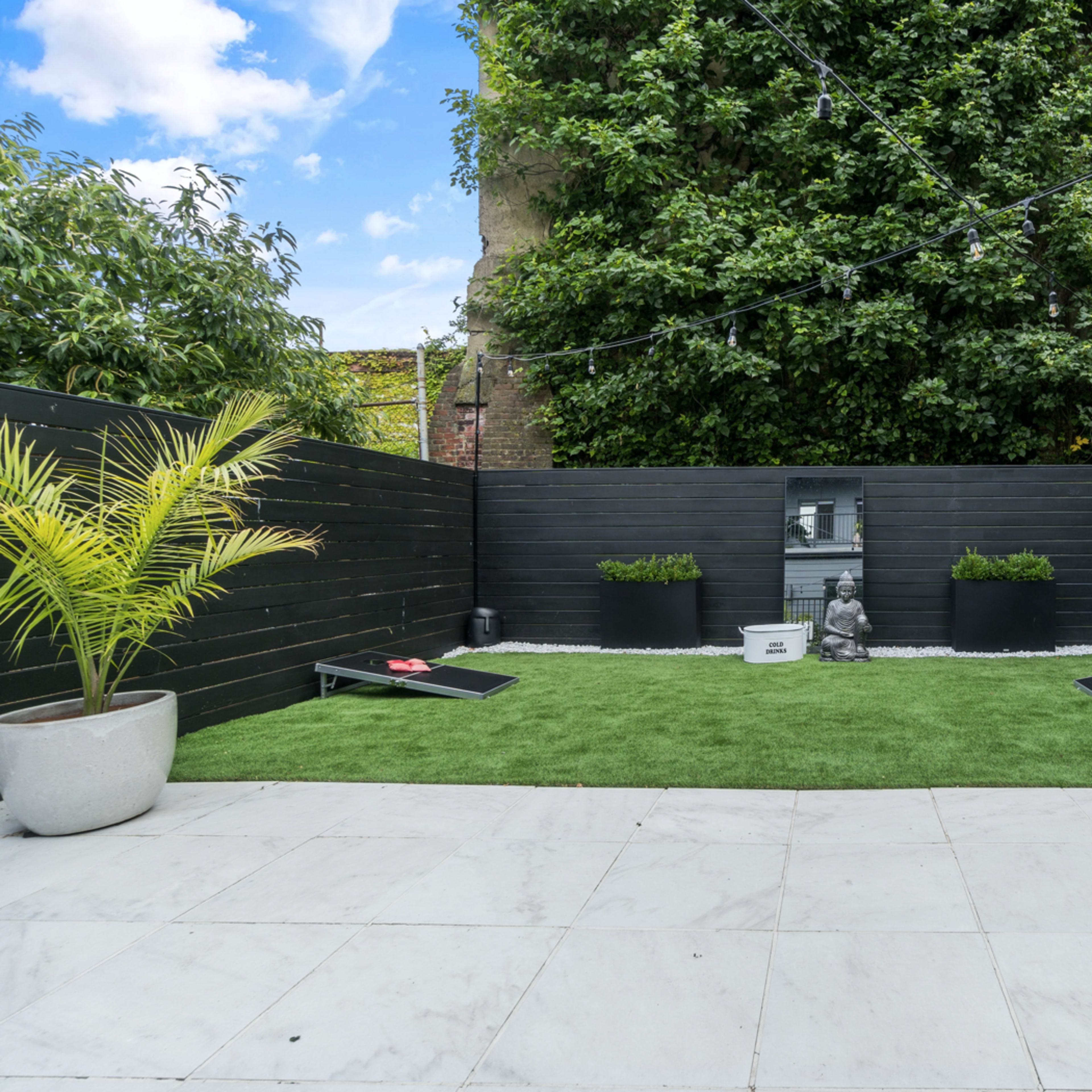 A compact garden with a grass area surrounded by black wooden fencing, potted plants, and a decorative statue.