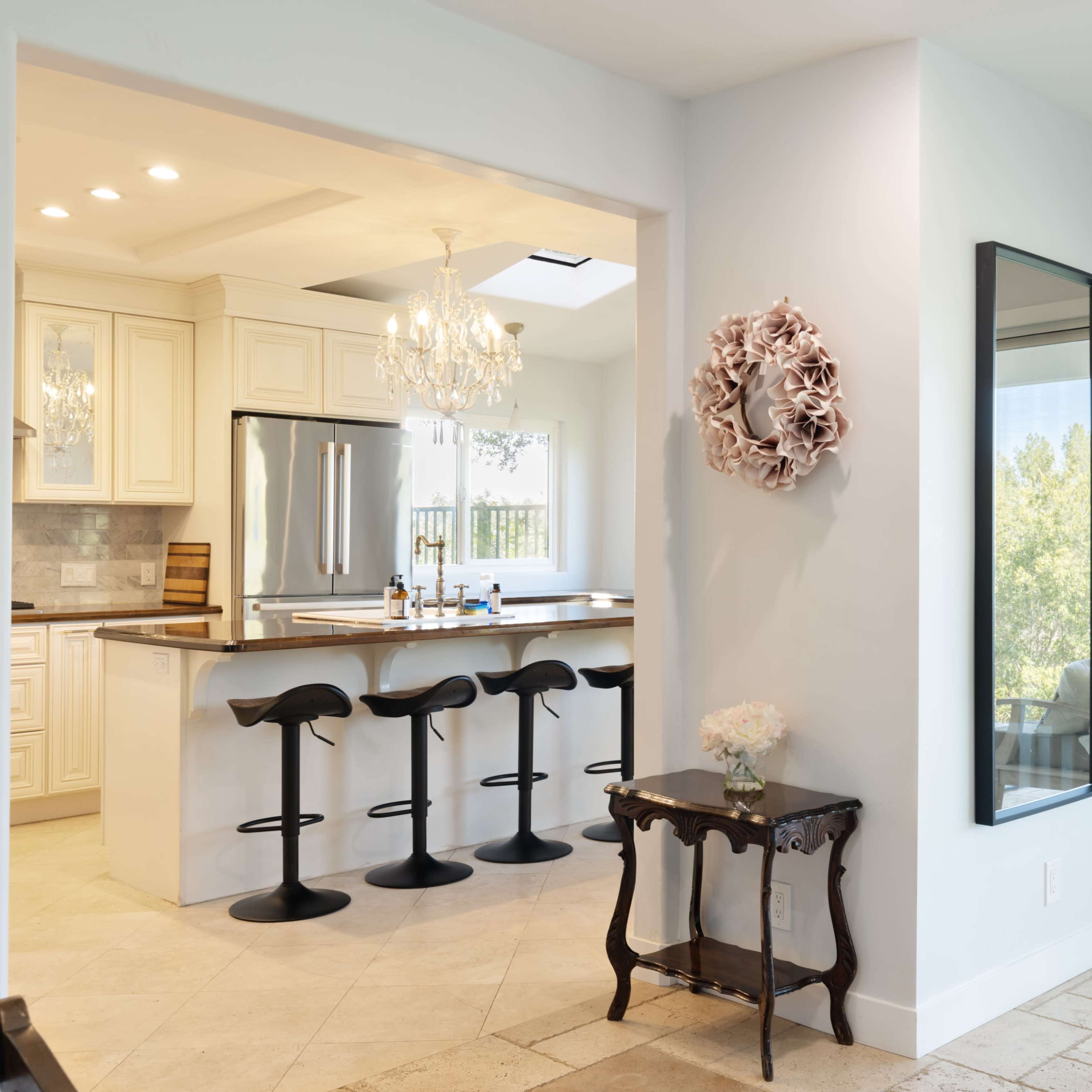 A modern kitchen visible from an adjacent living area, featuring a bar with three stools, a dining table, and large windows leading to an outdoor space.