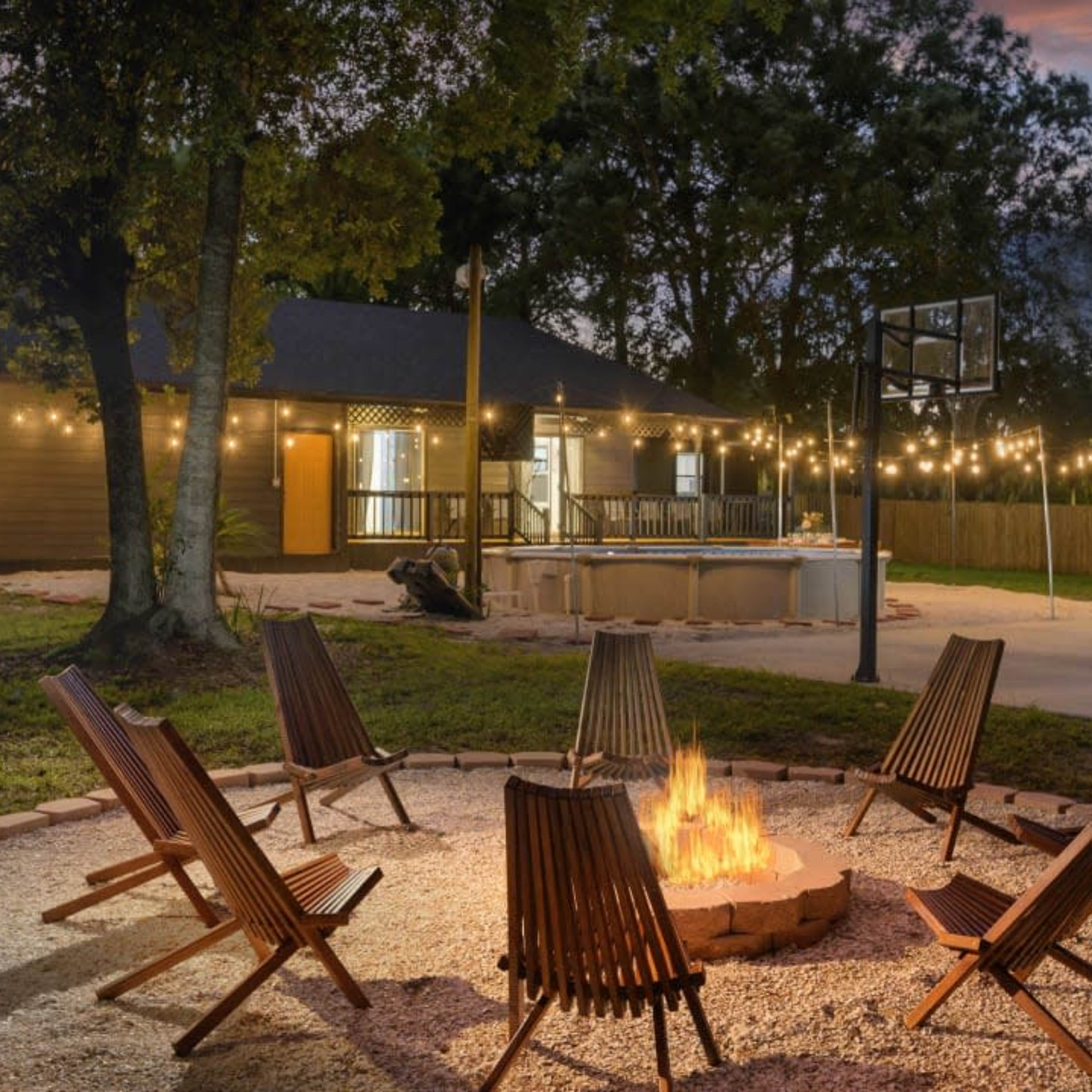 A circular fire pit surrounded by wooden chairs is situated in a backyard with string lights, a basketball hoop, and a house in the background.