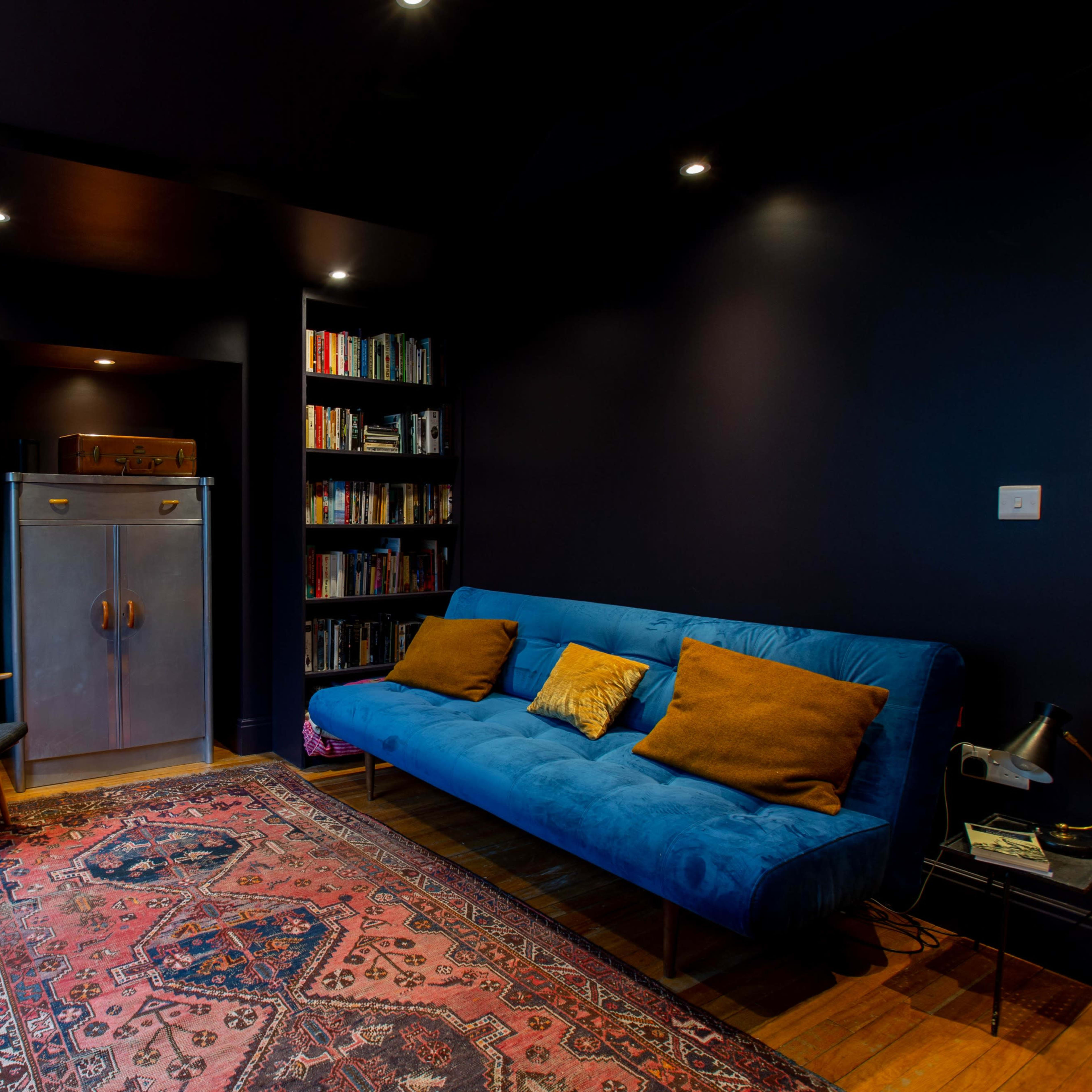 A dimly lit room features a blue sofa with decorative pillows, a wooden armchair, a bookshelf filled with books, and an antique cabinet against a dark wall.