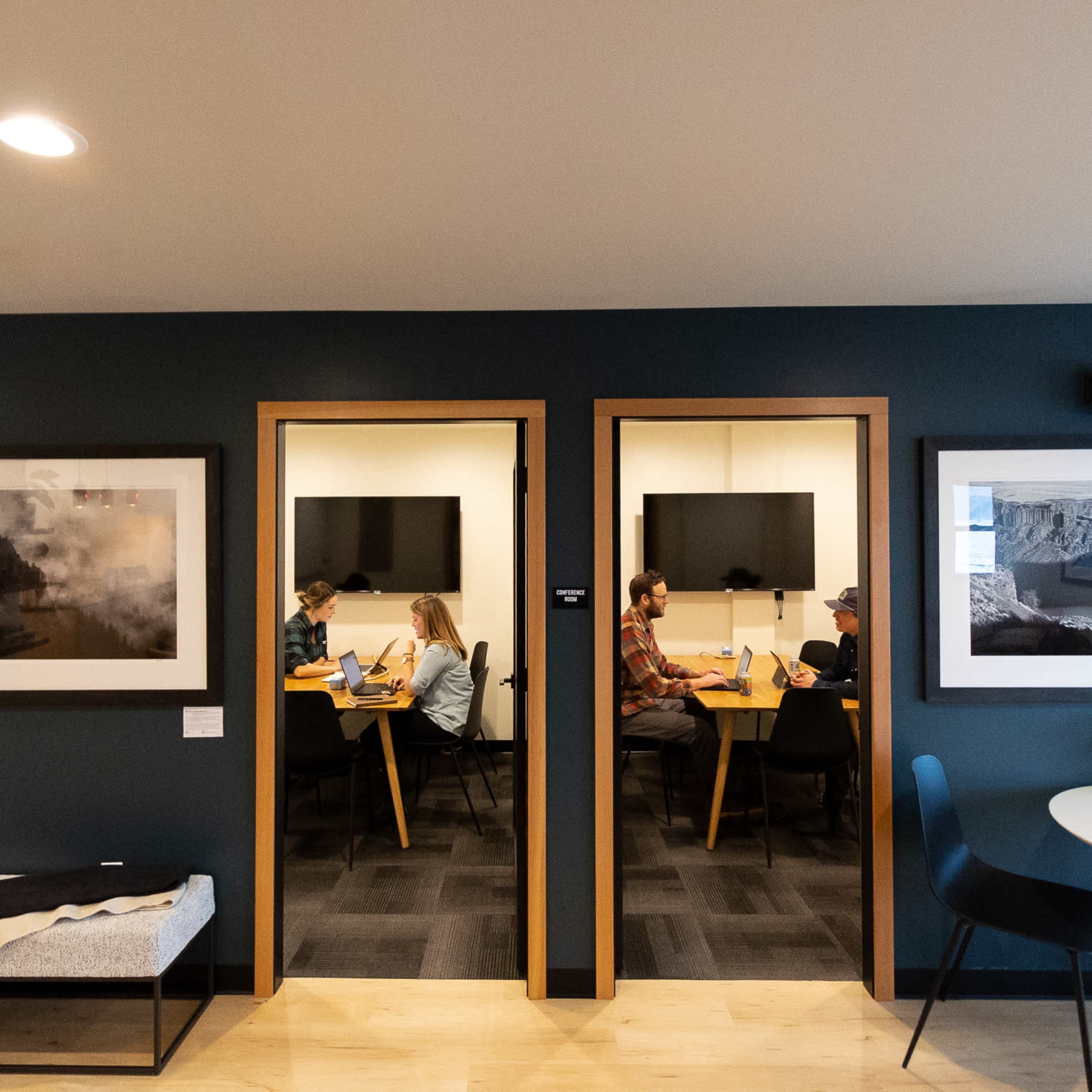 Two adjacent conference rooms with people seated at tables, each room equipped with a television and framed photographs on the walls.