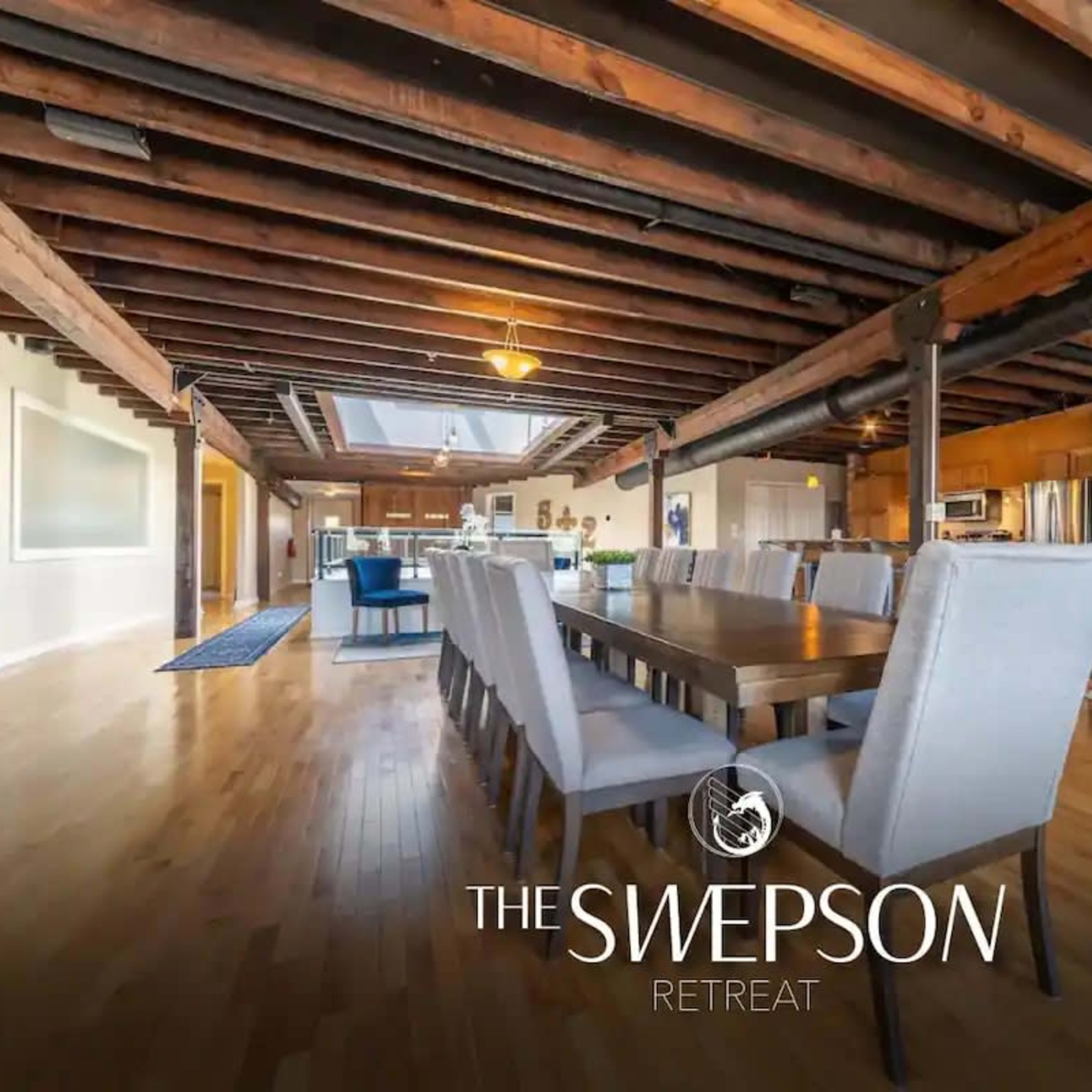 A spacious open-concept living area featuring a long dining table, wooden beams, and large windows that fill the room with natural light.