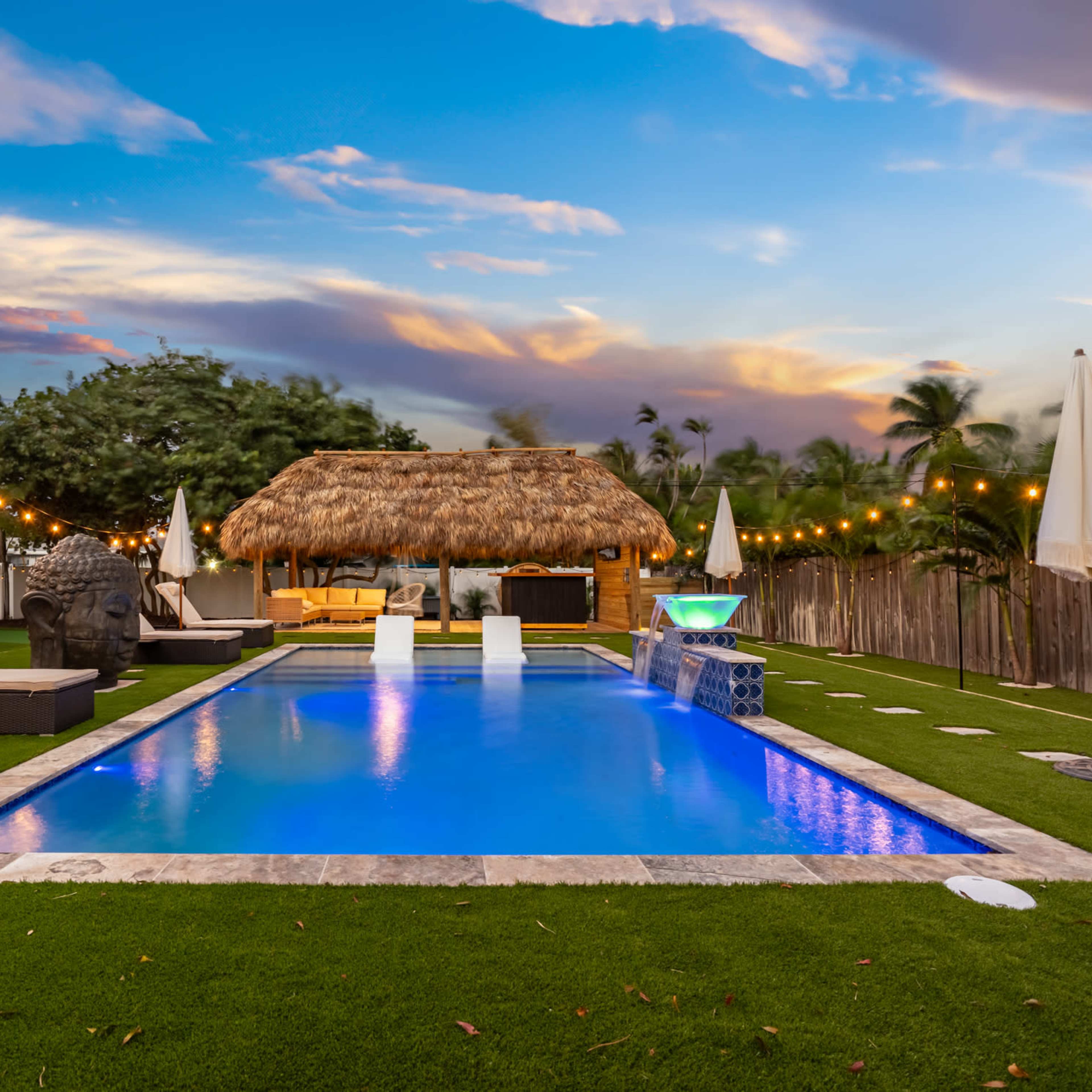 A swimming pool surrounded by lounge chairs and a thatched-roof gazebo is set in a green lawn under a colorful sky.