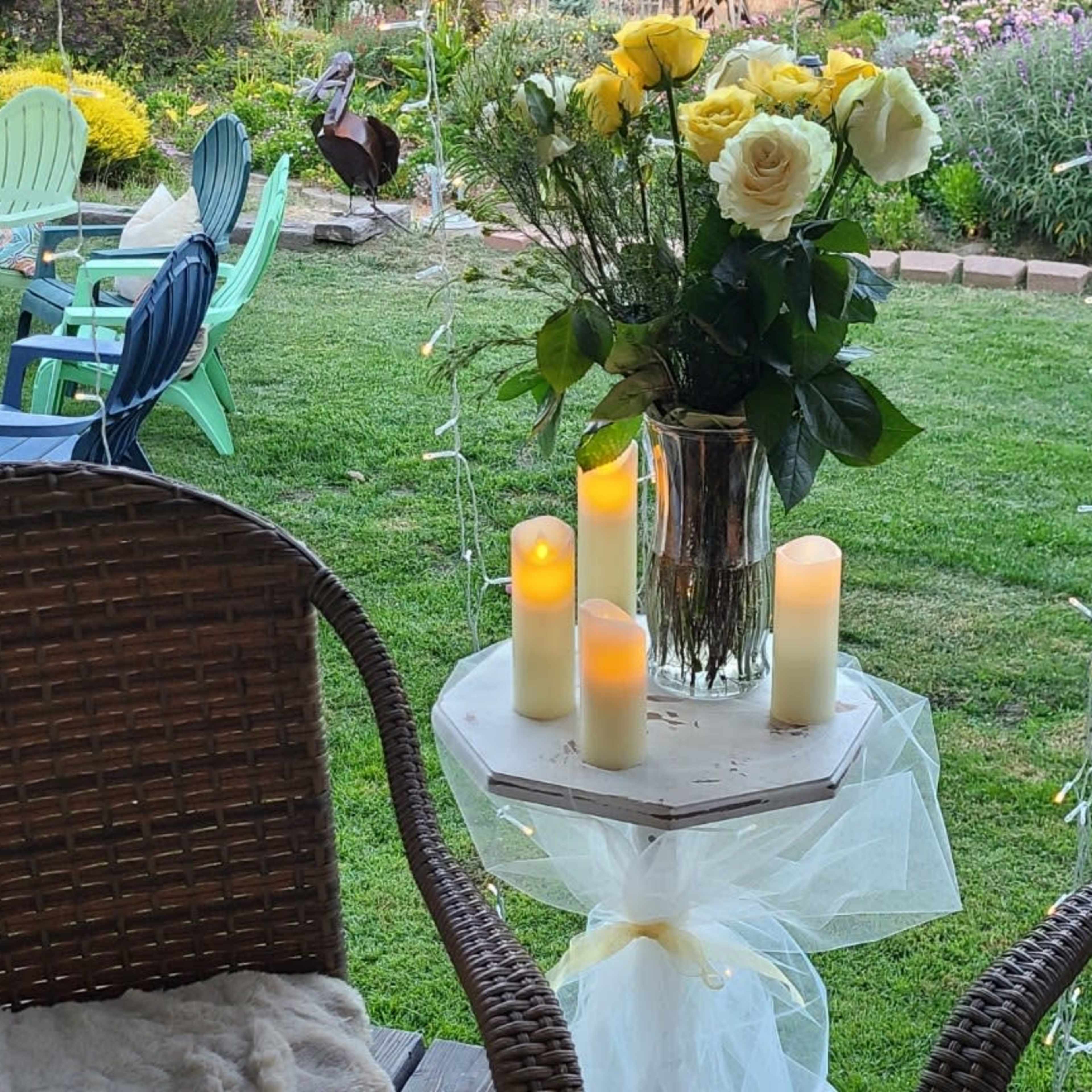 A table with a vase of yellow and white roses and candles stands between two wicker chairs in a garden setting.