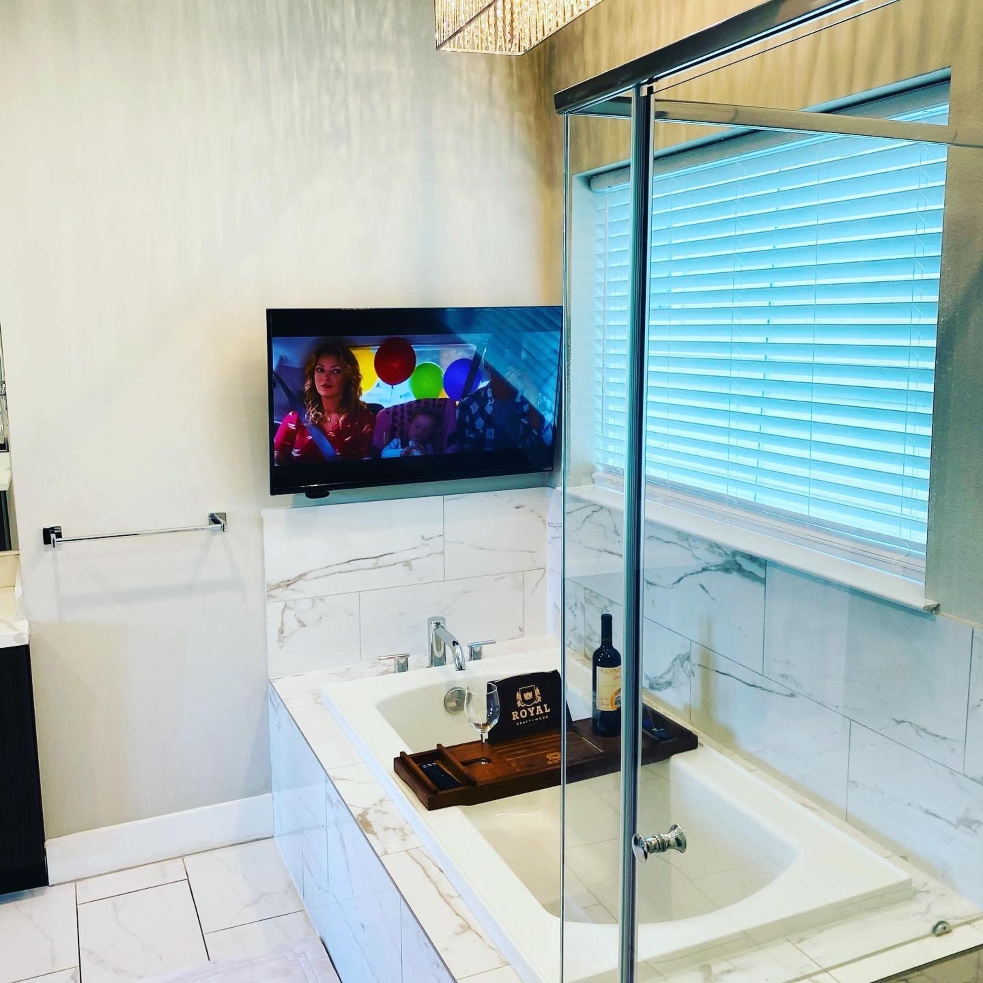 A modern bathroom features a bathtub with a wooden tray, a television mounted on the wall playing a show, and a window with blinds providing natural light.