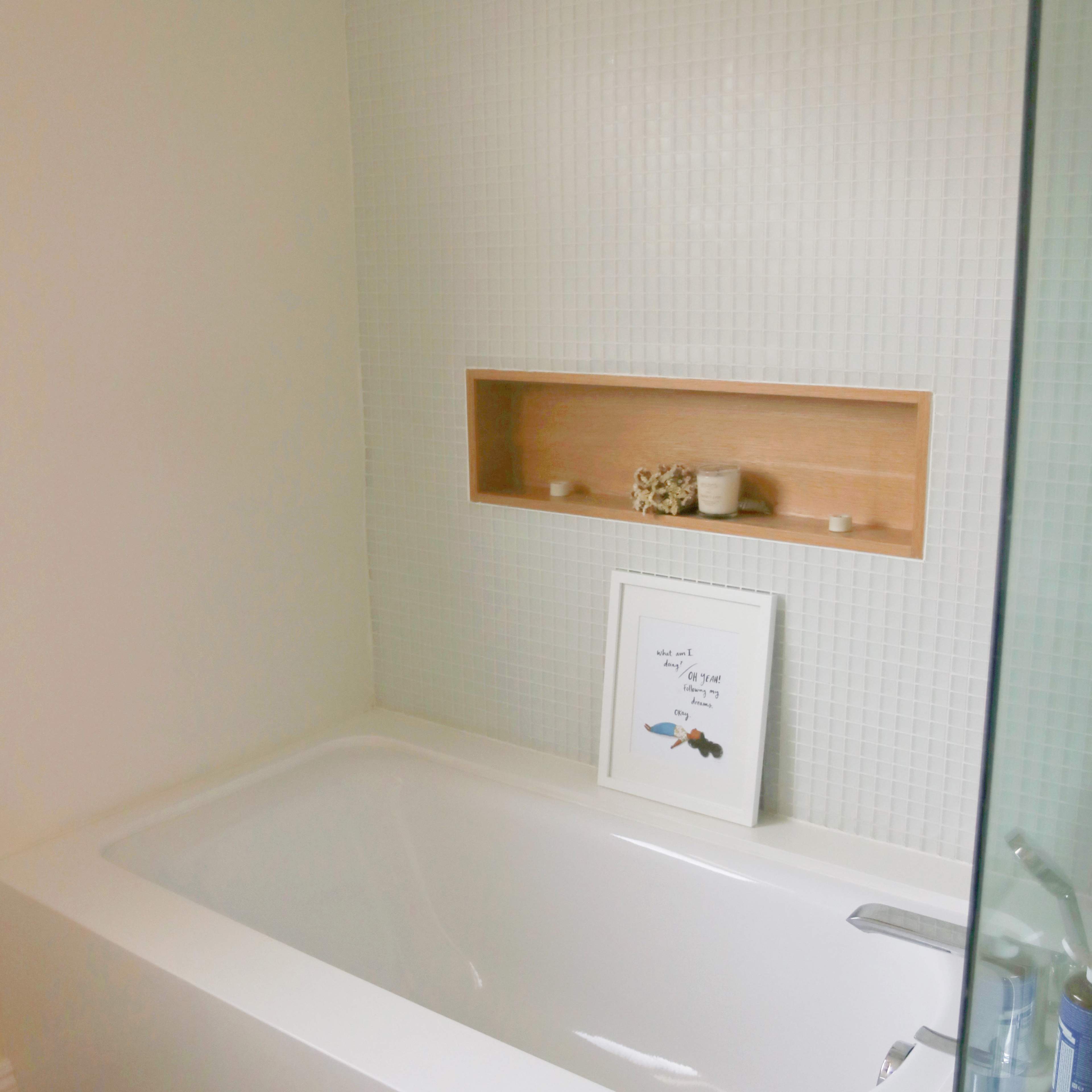 A clean and modern bathroom features a white bathtub, a wooden shelf with decorative items, and tiled walls.
