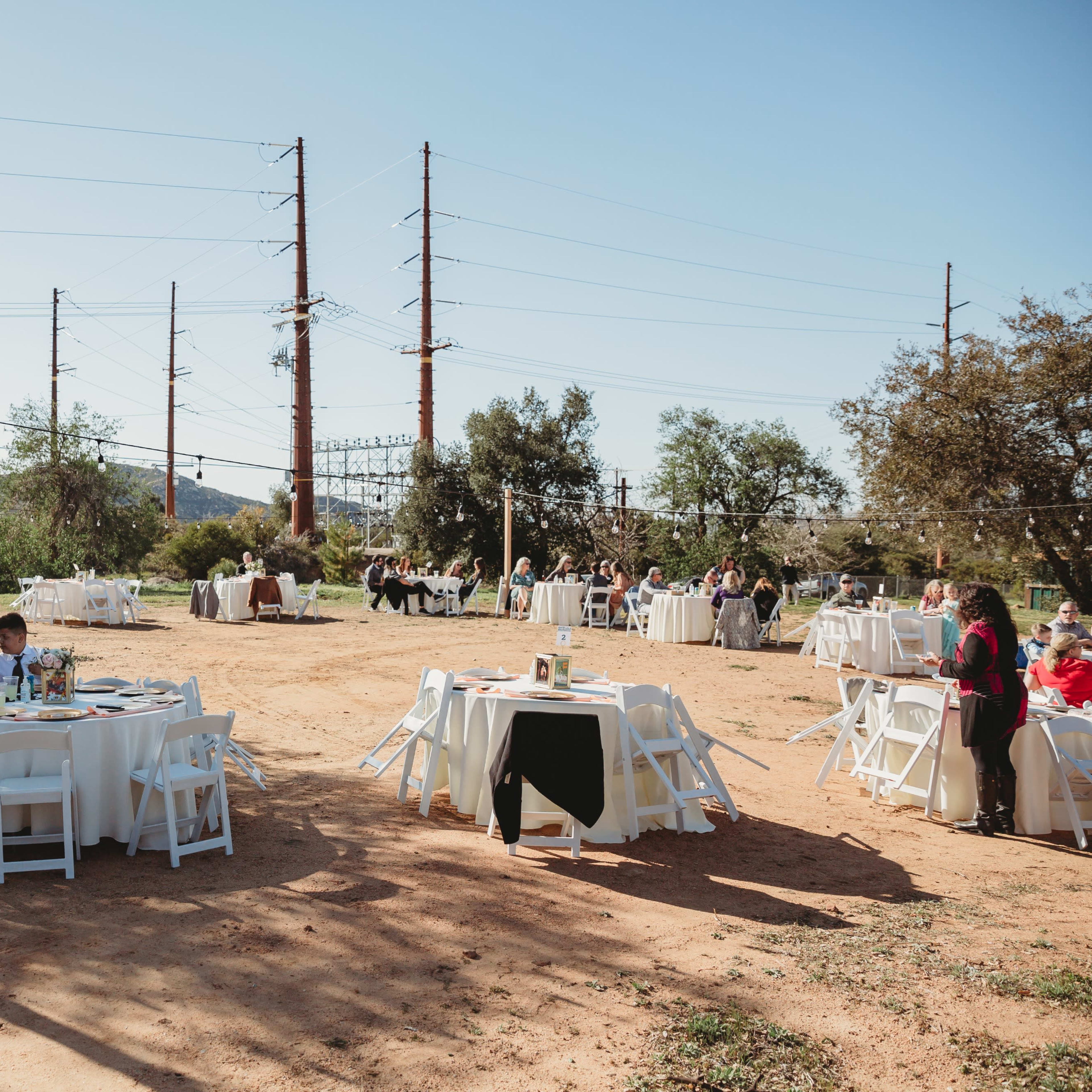 A reception area with several round tables set up outdoors, surrounded by trees and power lines, hosts guests enjoying a meal.