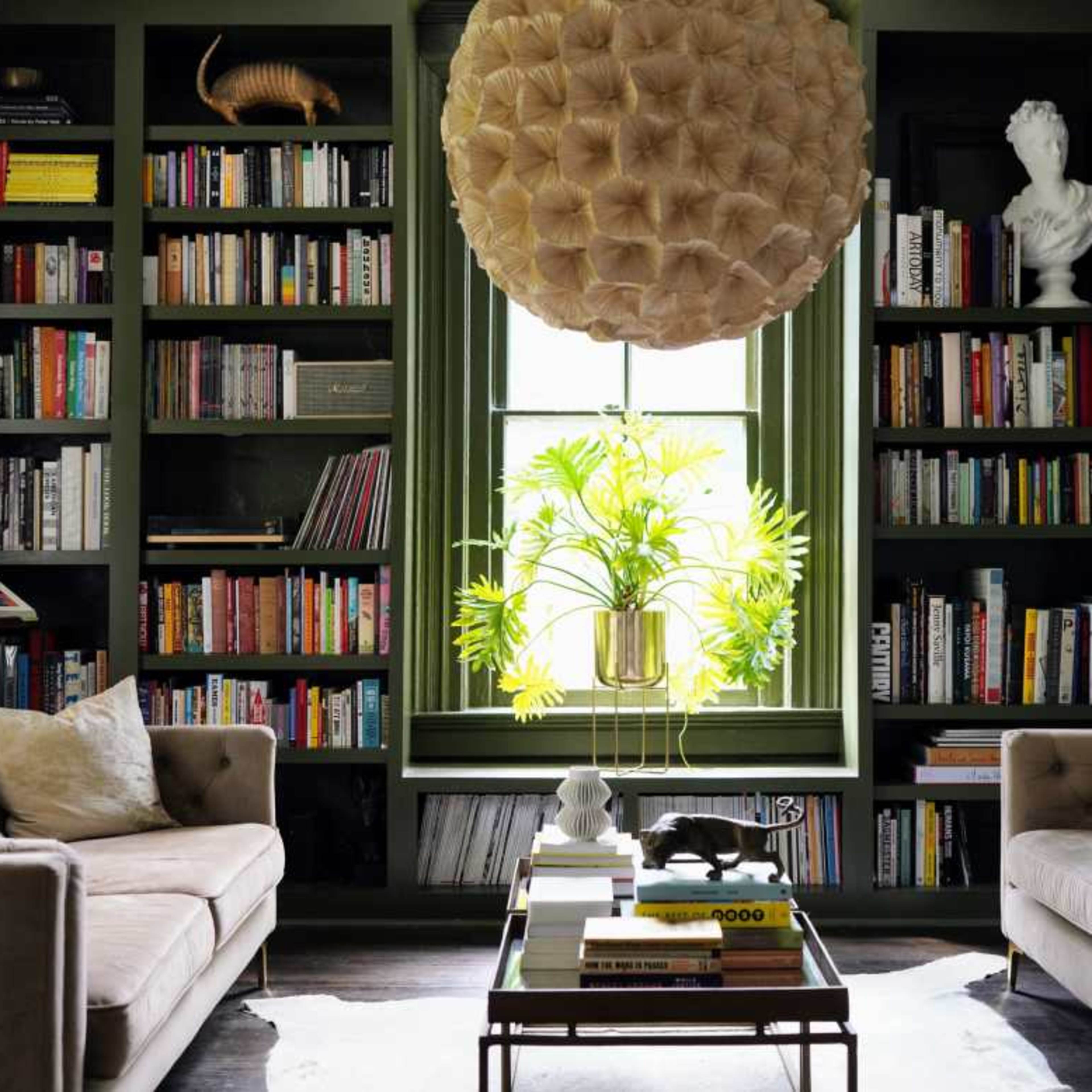 A cozy living room features two sofas facing a coffee table, surrounded by tall bookshelves filled with books and adorned with a large pendant light and decorative plants.