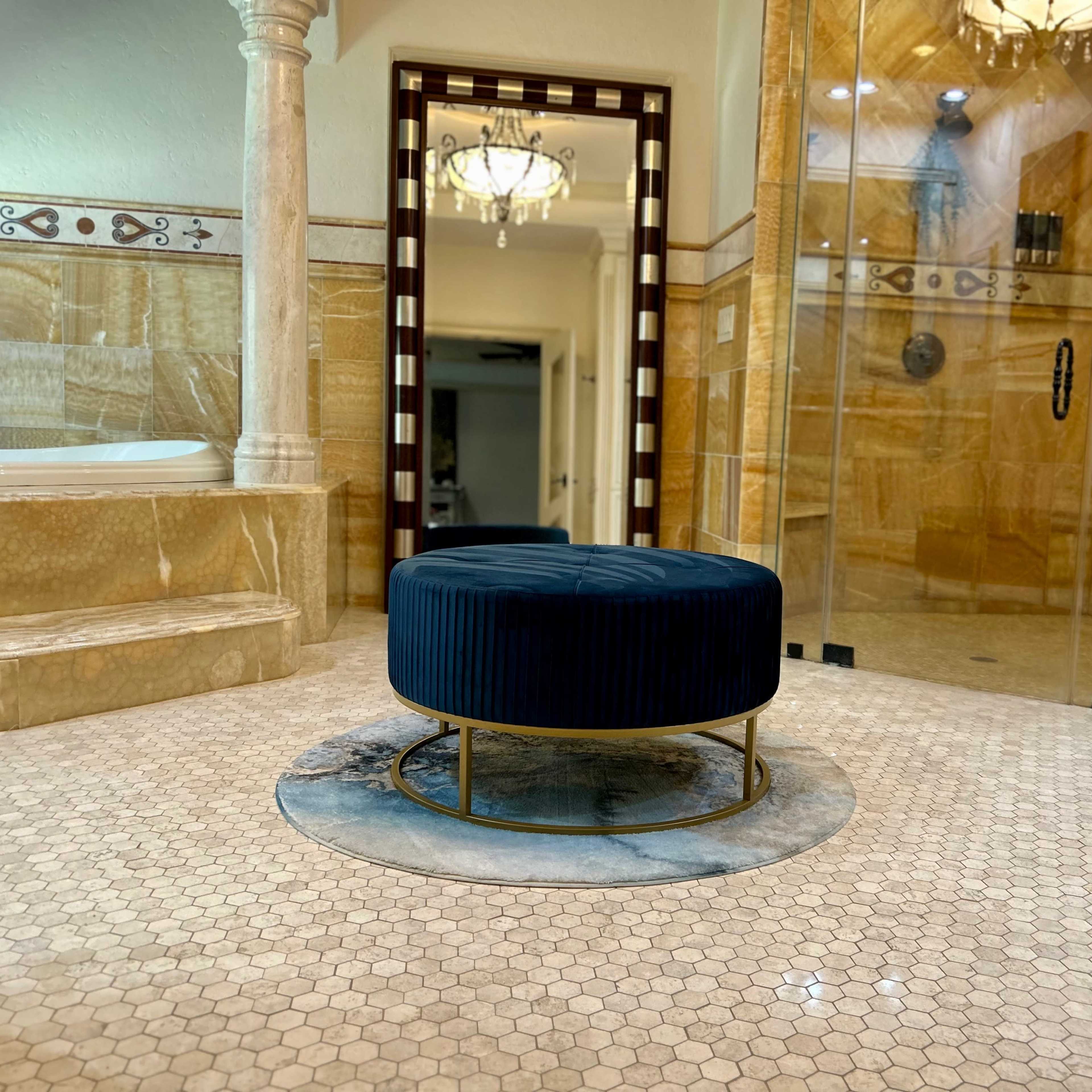 A luxurious bathroom featuring a round navy ottoman centrally placed on a hexagonal tile floor, with a marble rug, a large mirror, and a walk-in shower in the background.