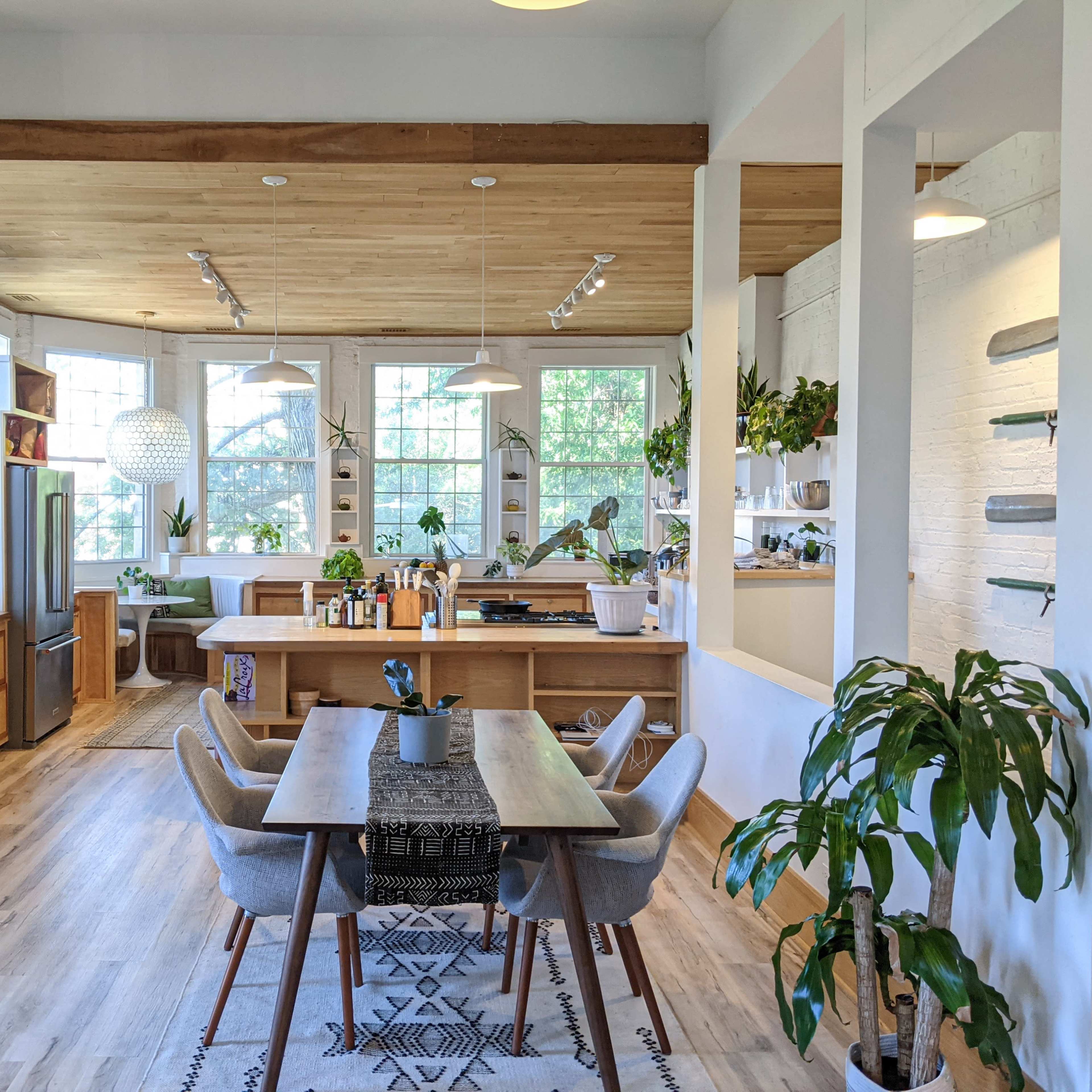 A bright, modern kitchen and dining area featuring wooden cabinetry, a large dining table, and numerous houseplants.