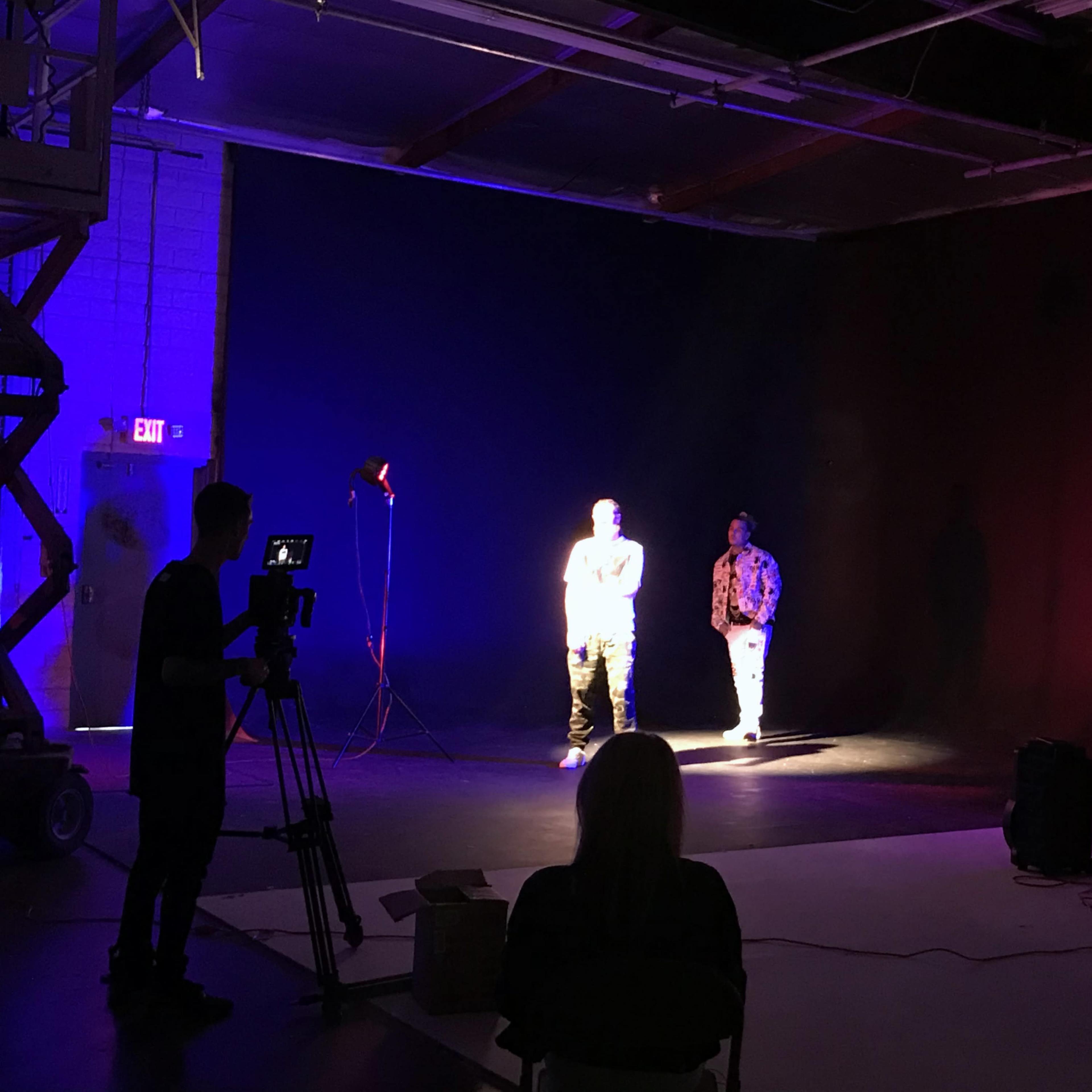 Two individuals are illuminated in a dimly lit studio, surrounded by various stage lights and camera equipment.