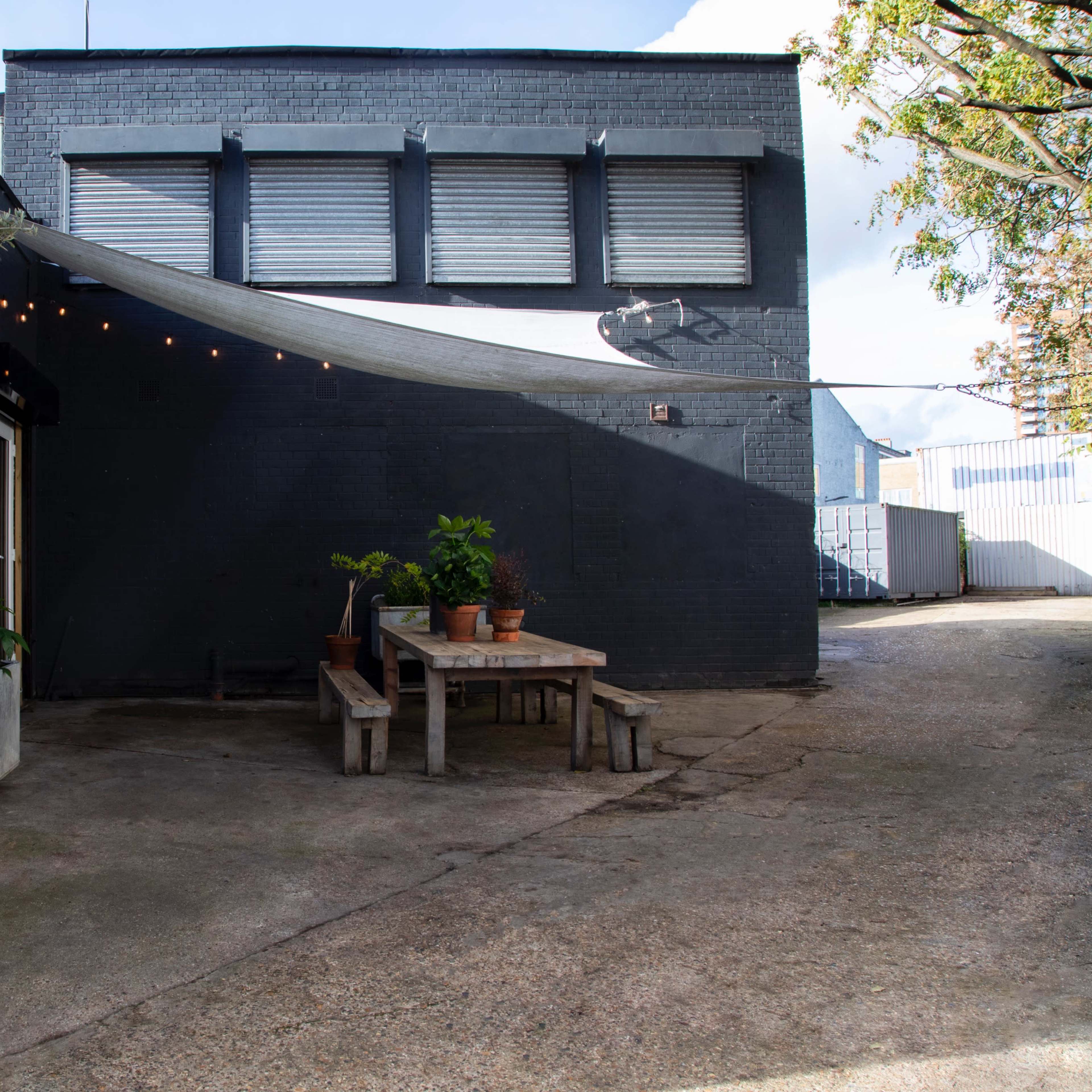 A gray wall of a building with a wooden table and benches, surrounded by potted plants and an olive tree, under a canopy of string lights.