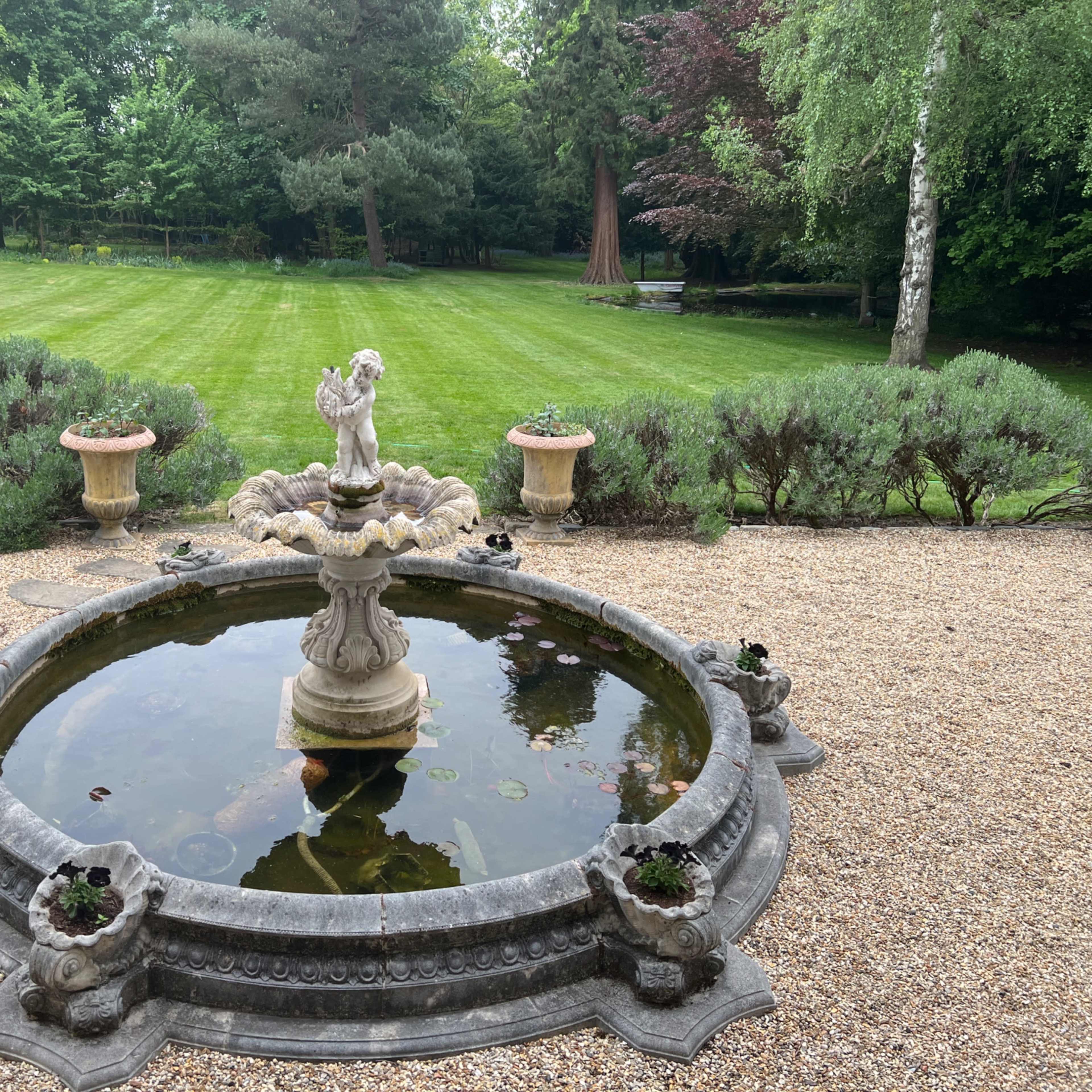 A stone fountain with a statue of an angel is surrounded by a circular pond in a garden with manicured grass and shrubs.