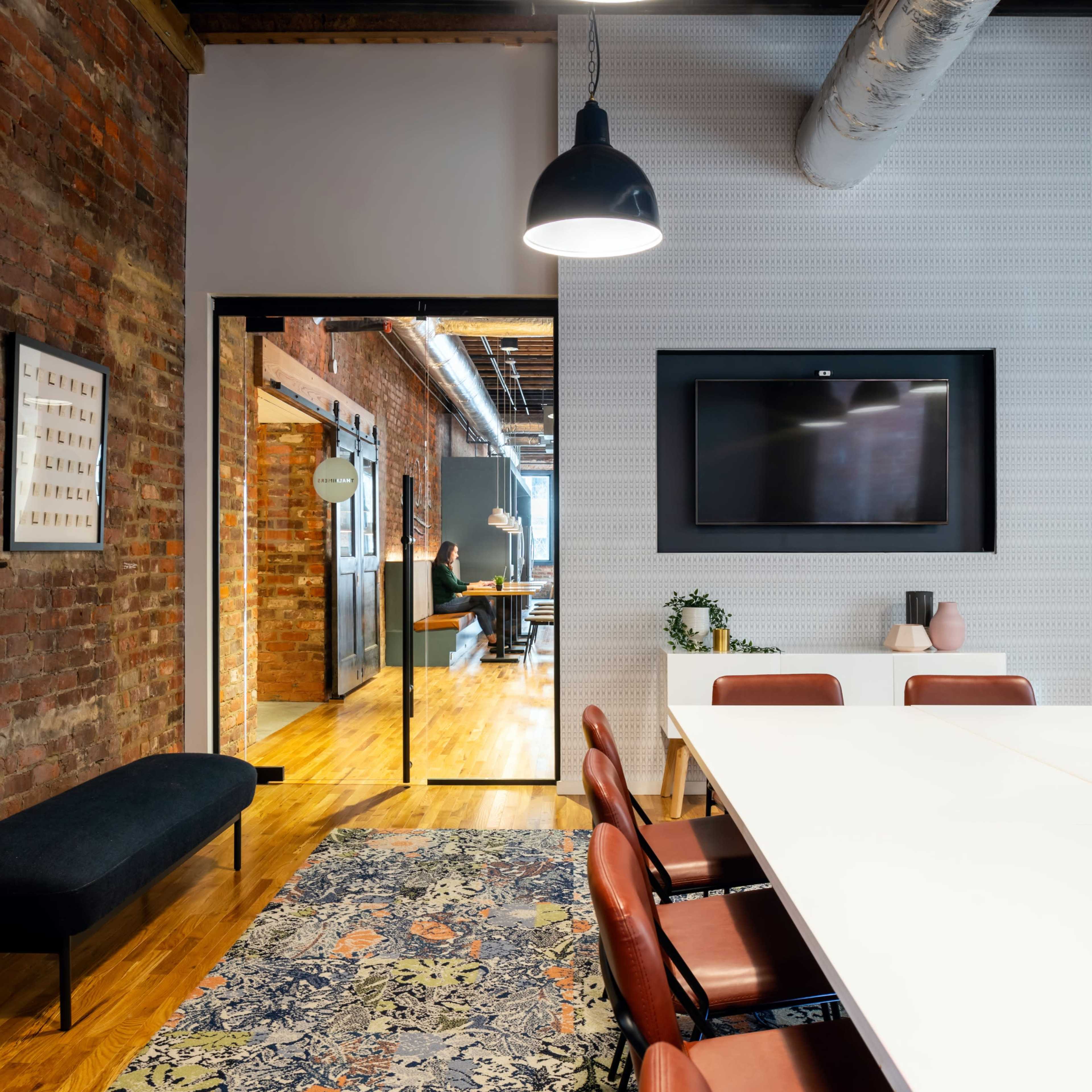 A modern conference room features a long white table surrounded by brown chairs, with exposed brick walls, a television mounted on the wall, and a decorative plant in the corner.