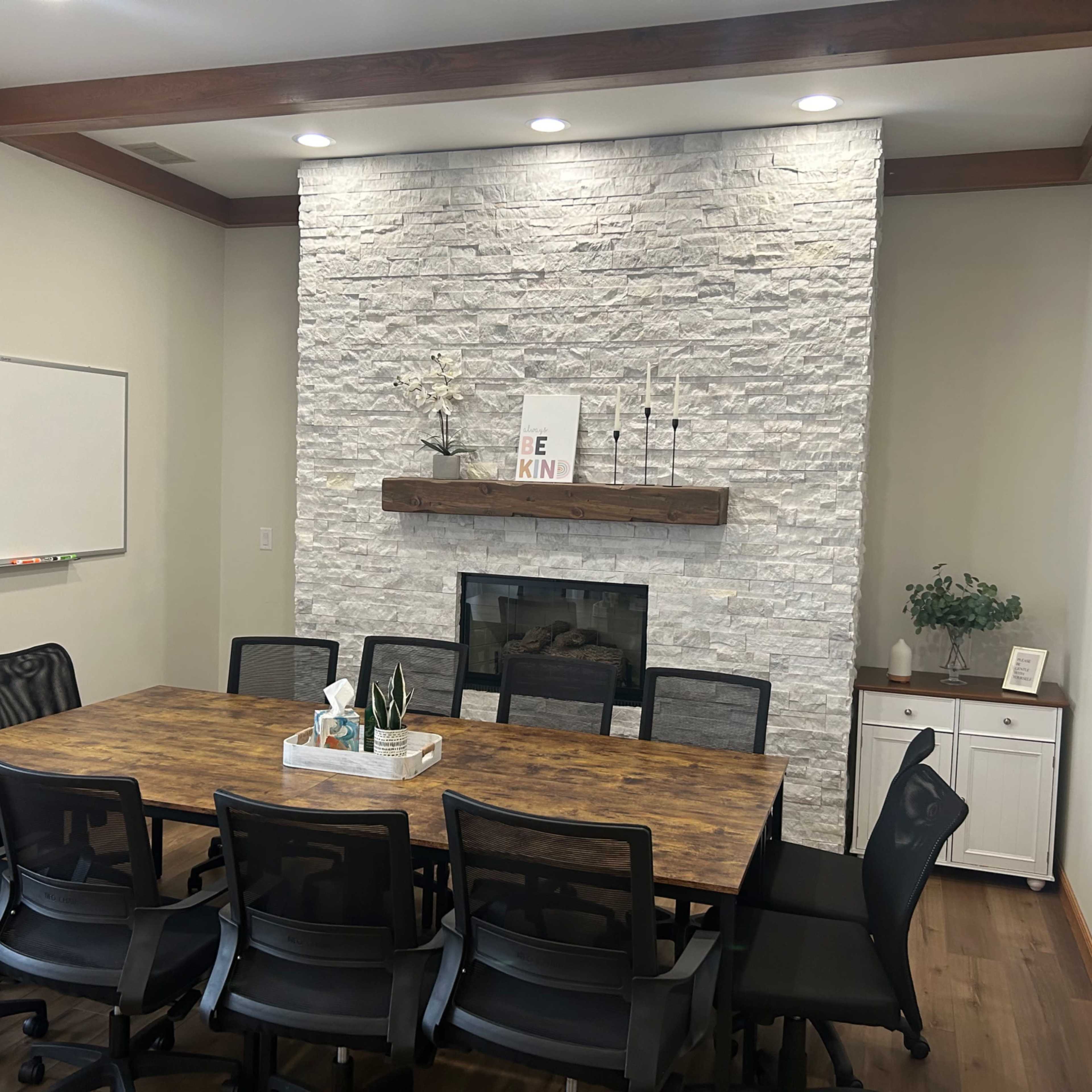 A conference room features a large wooden table surrounded by black chairs, a stone fireplace with a mantel, and whiteboards on the walls.