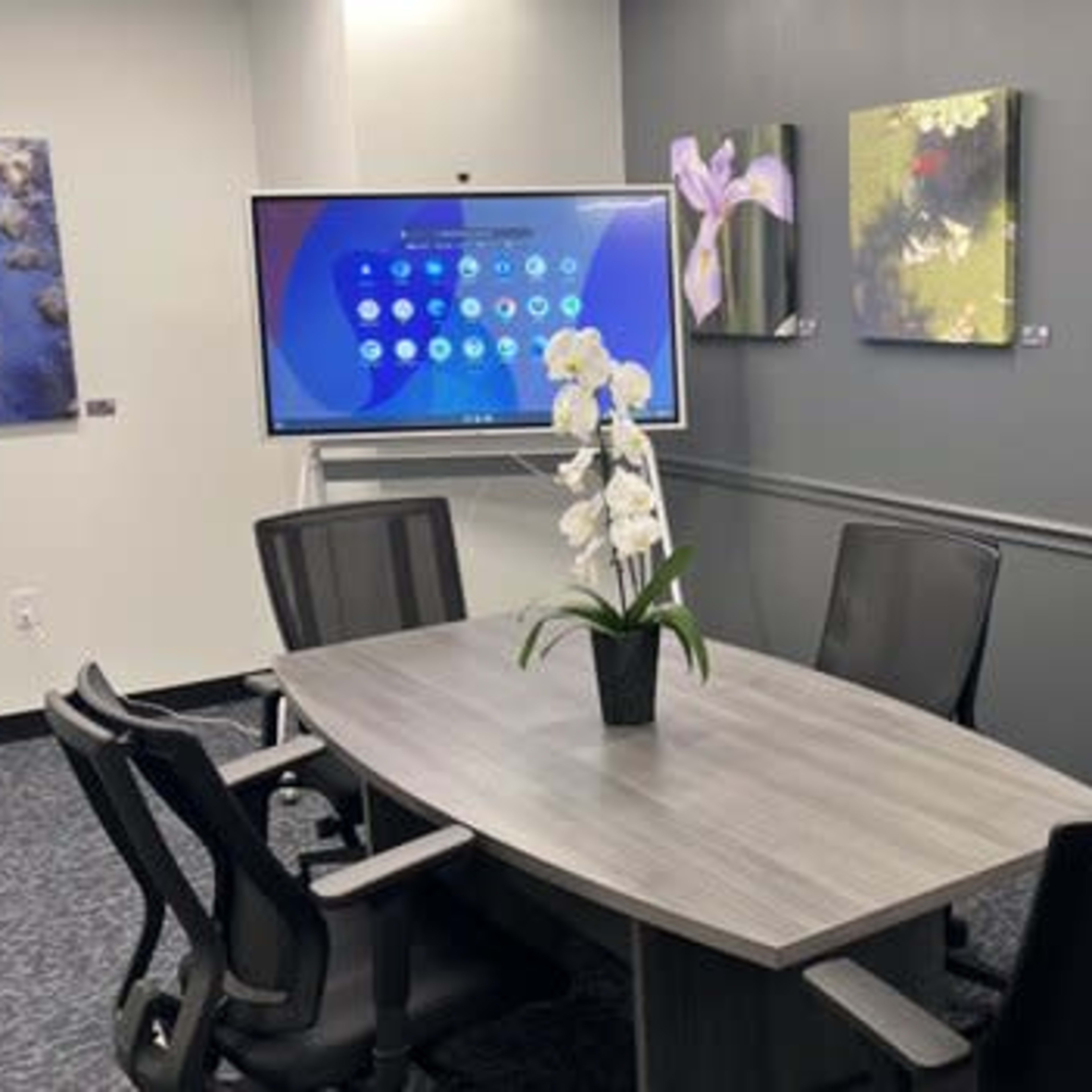 A modern conference room features a rectangular table surrounded by four chairs, a large screen mounted on the wall, and framed floral artwork displayed nearby.