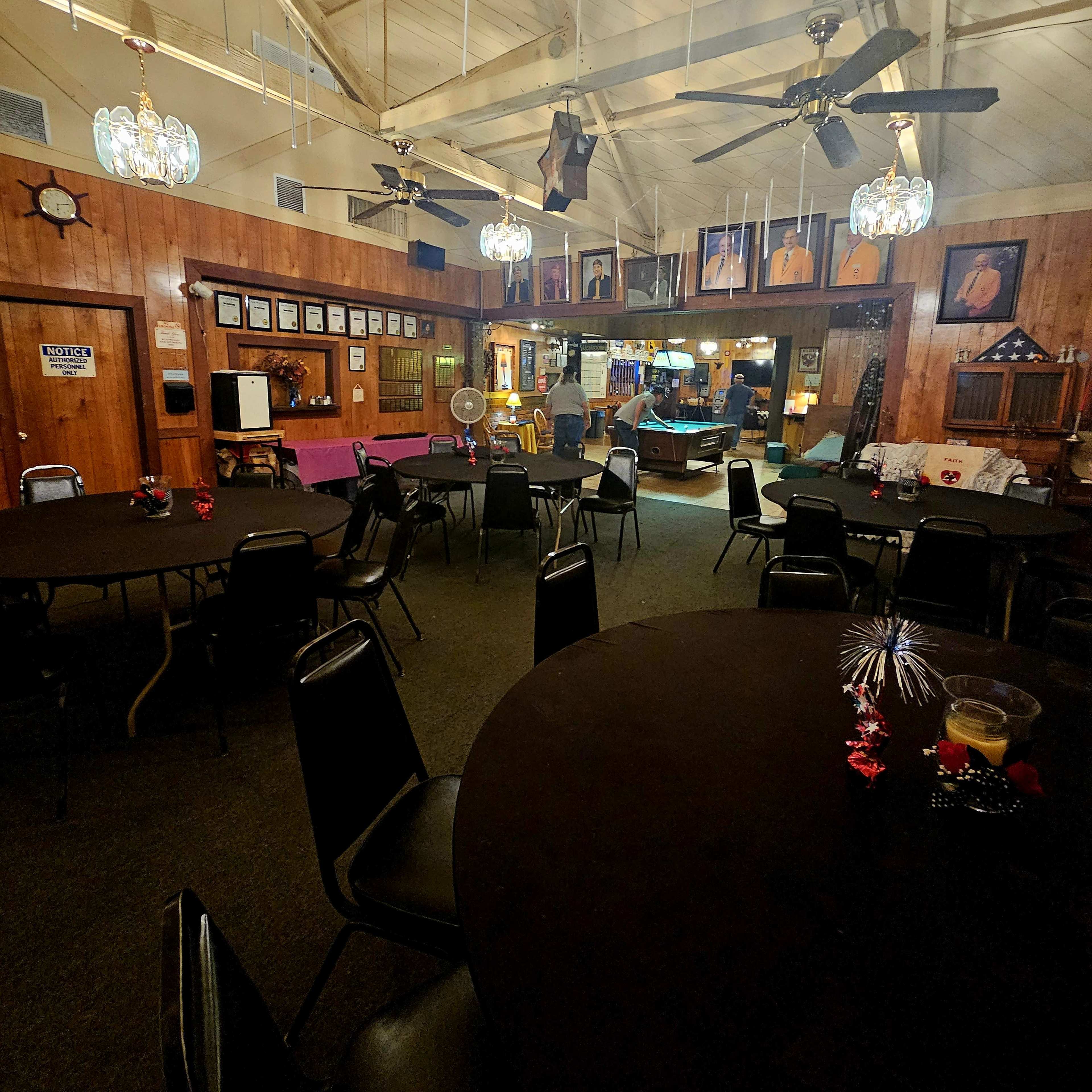 A spacious indoor setting with several round black tables and chairs, illuminated by decorative light fixtures and featuring a stage area in the background.