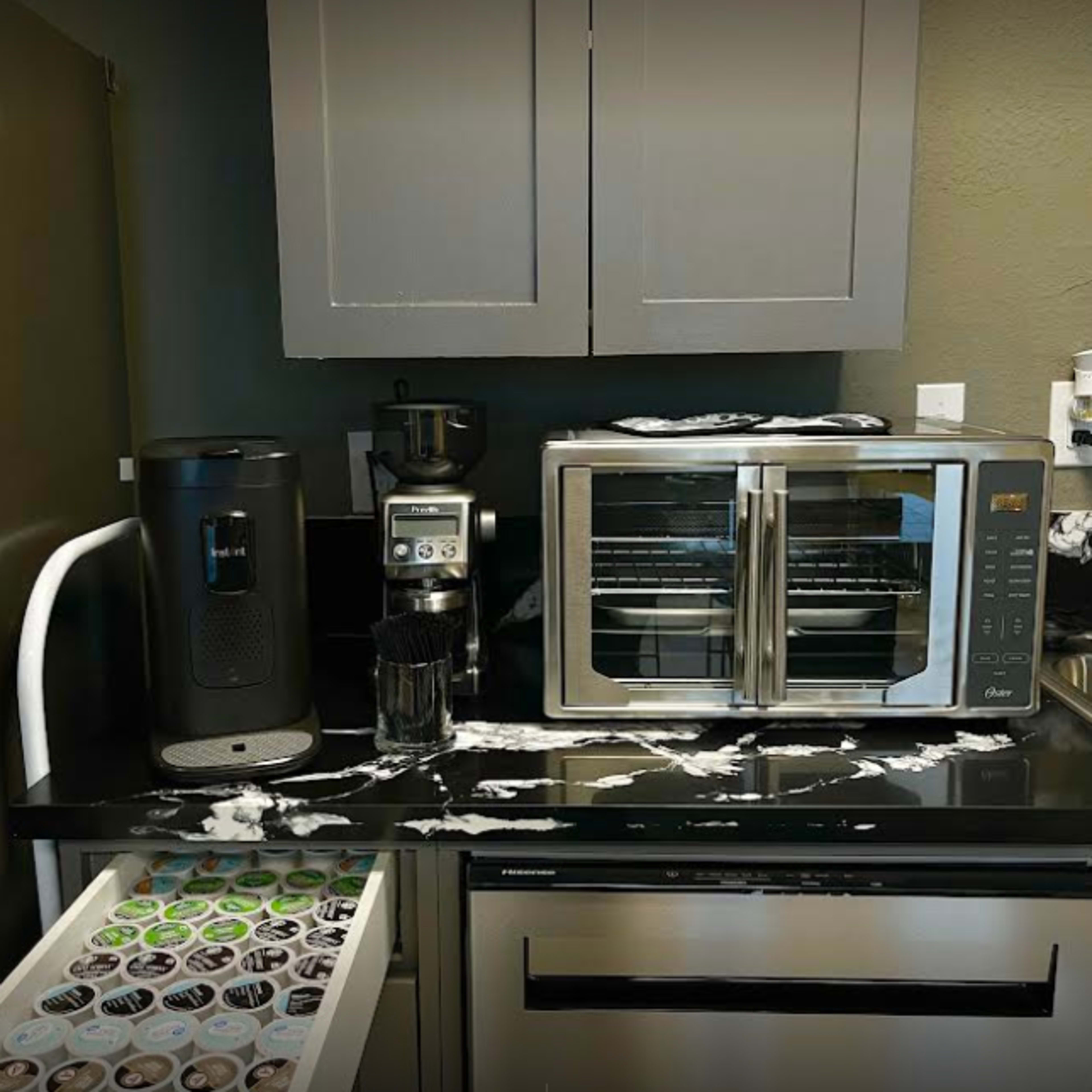 A kitchen countertop features a coffee maker, a toaster oven, and a drawer filled with coffee pods, with what appears to be spilled liquid across the surface.