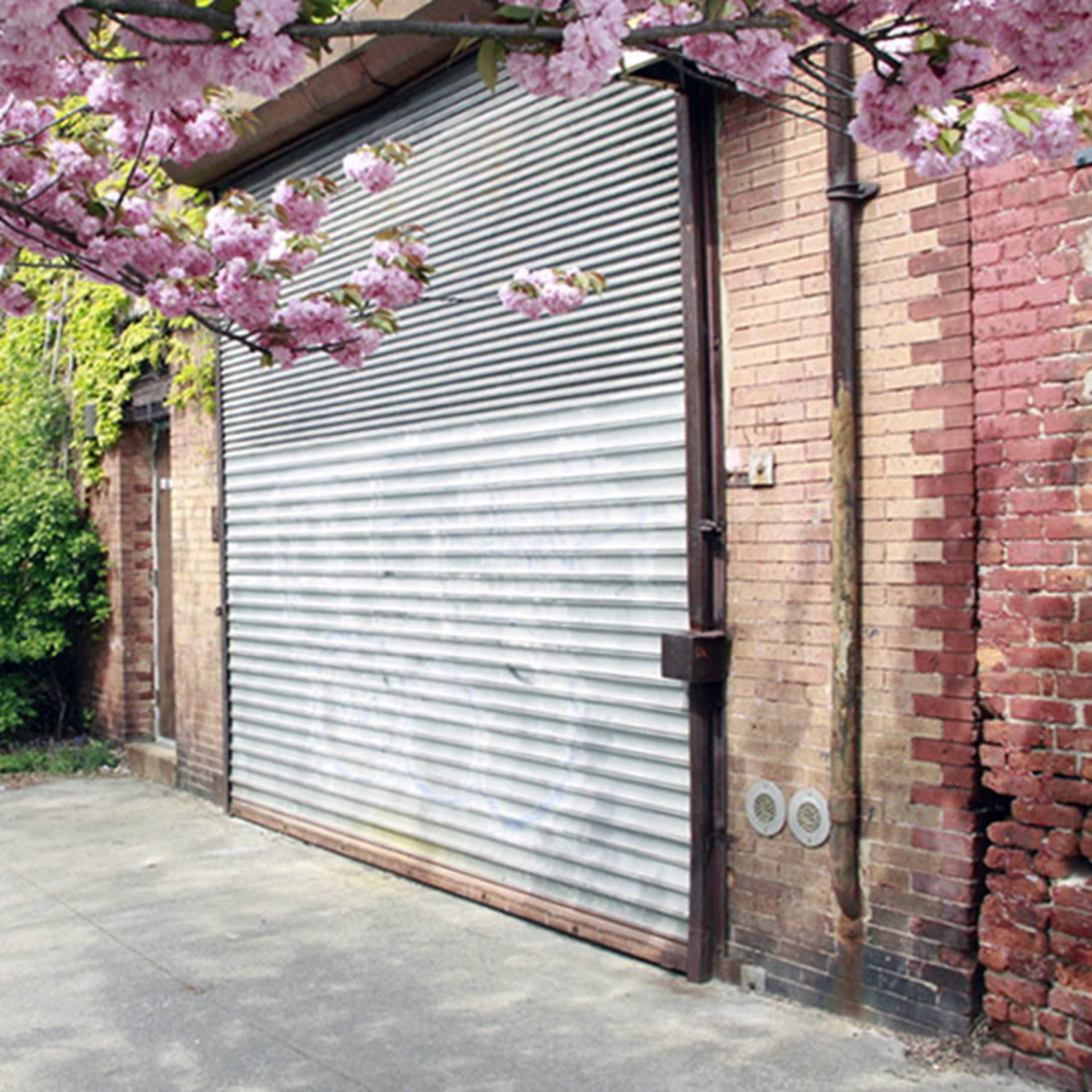 A person walks past a brick building with a closed metal garage door, framed by blooming cherry blossoms.