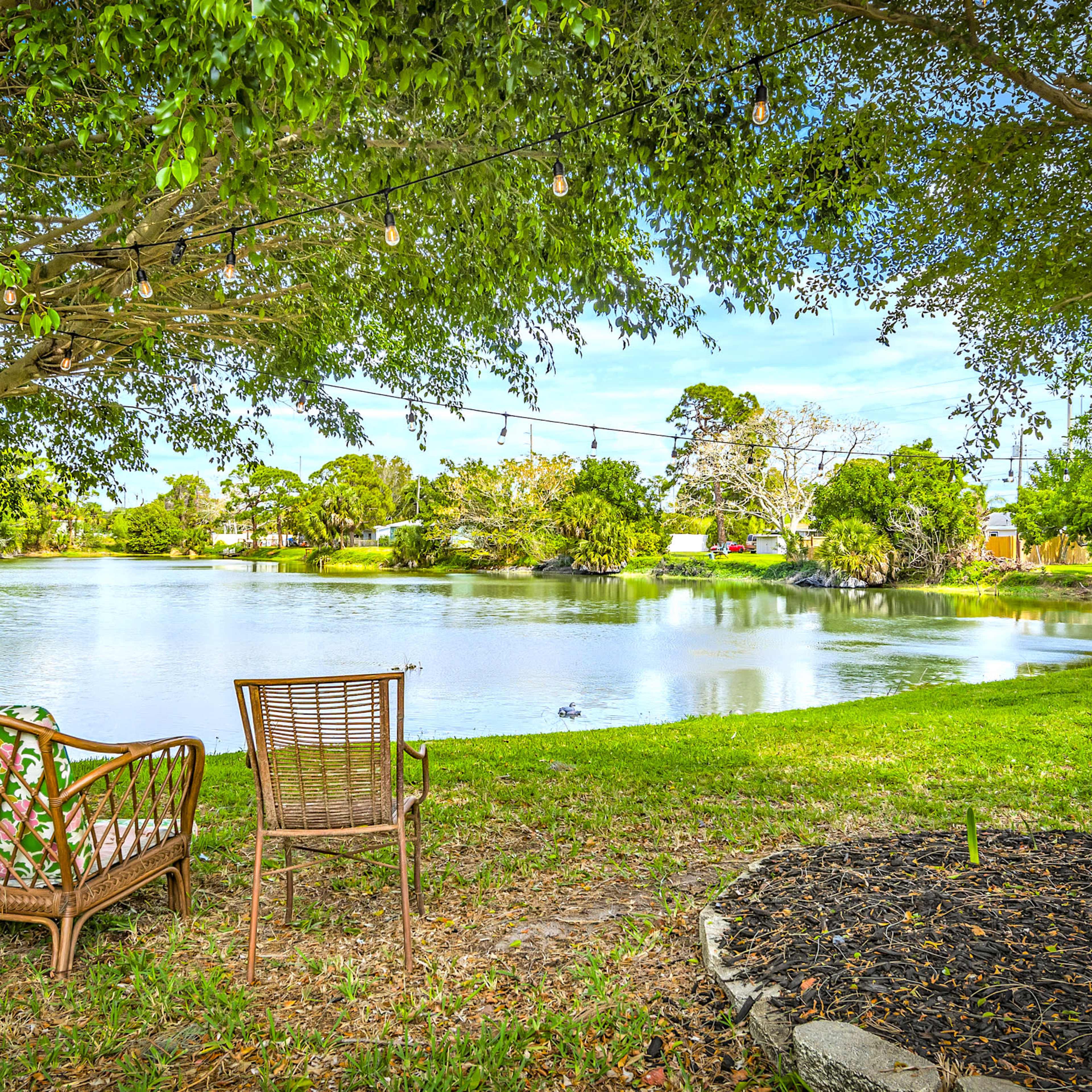 A pair of wicker chairs is positioned near a calm lake surrounded by trees and greenery, with decorative string lights overhead.