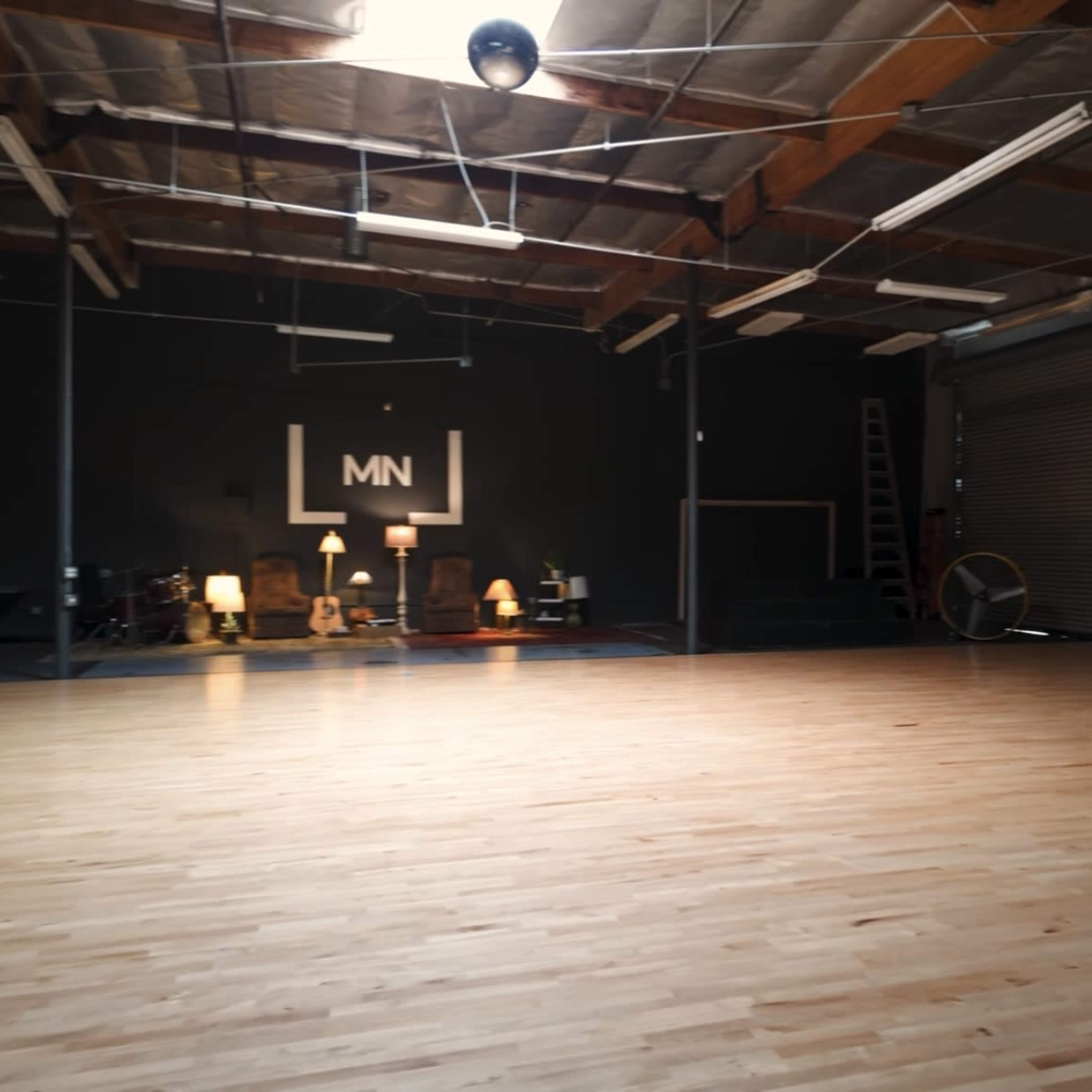 A spacious dance studio with wooden flooring, high ceilings, and minimal furnishings, illuminated by overhead lights.