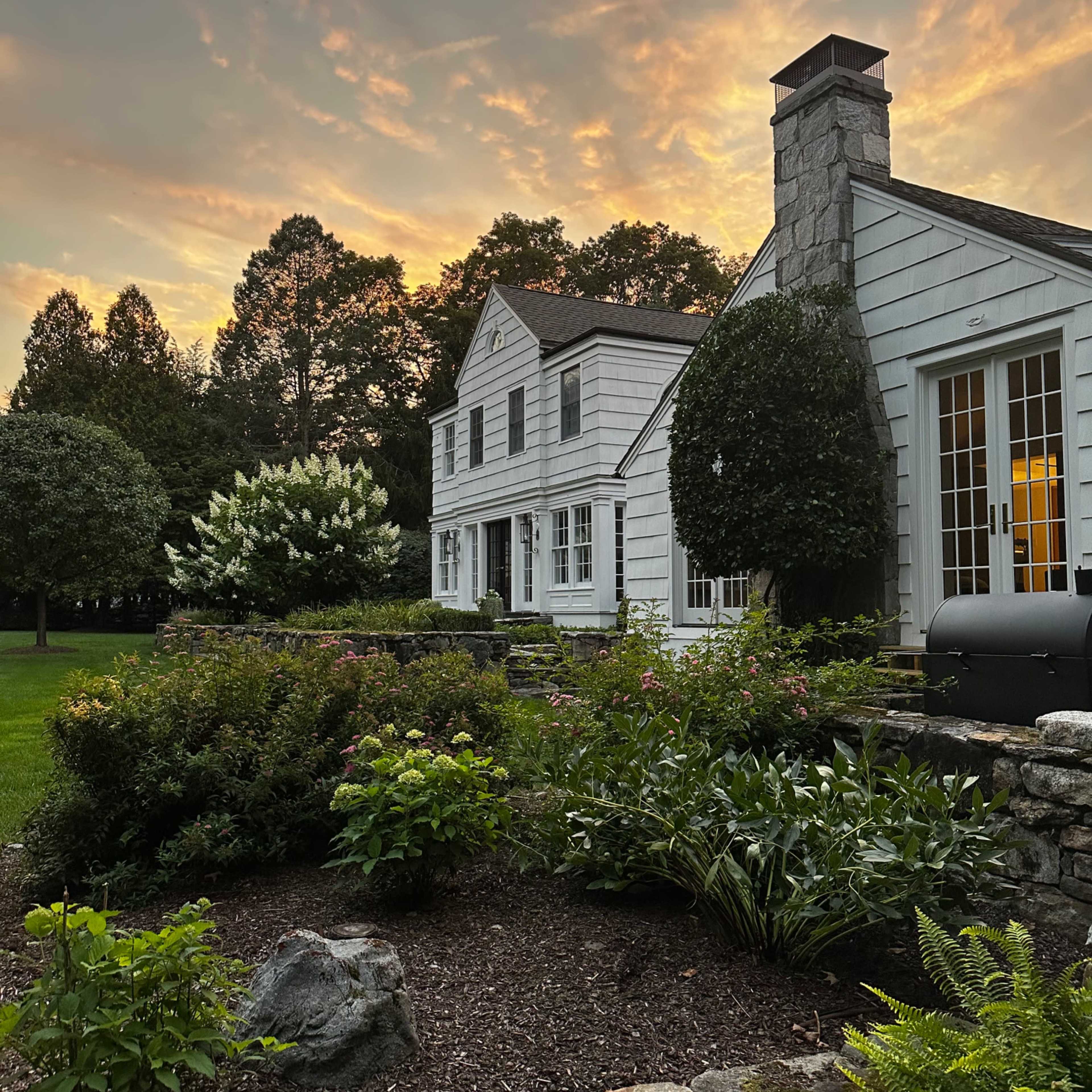 A white two-story house with a stone chimney is surrounded by well-maintained gardens under a colorful sunset sky.