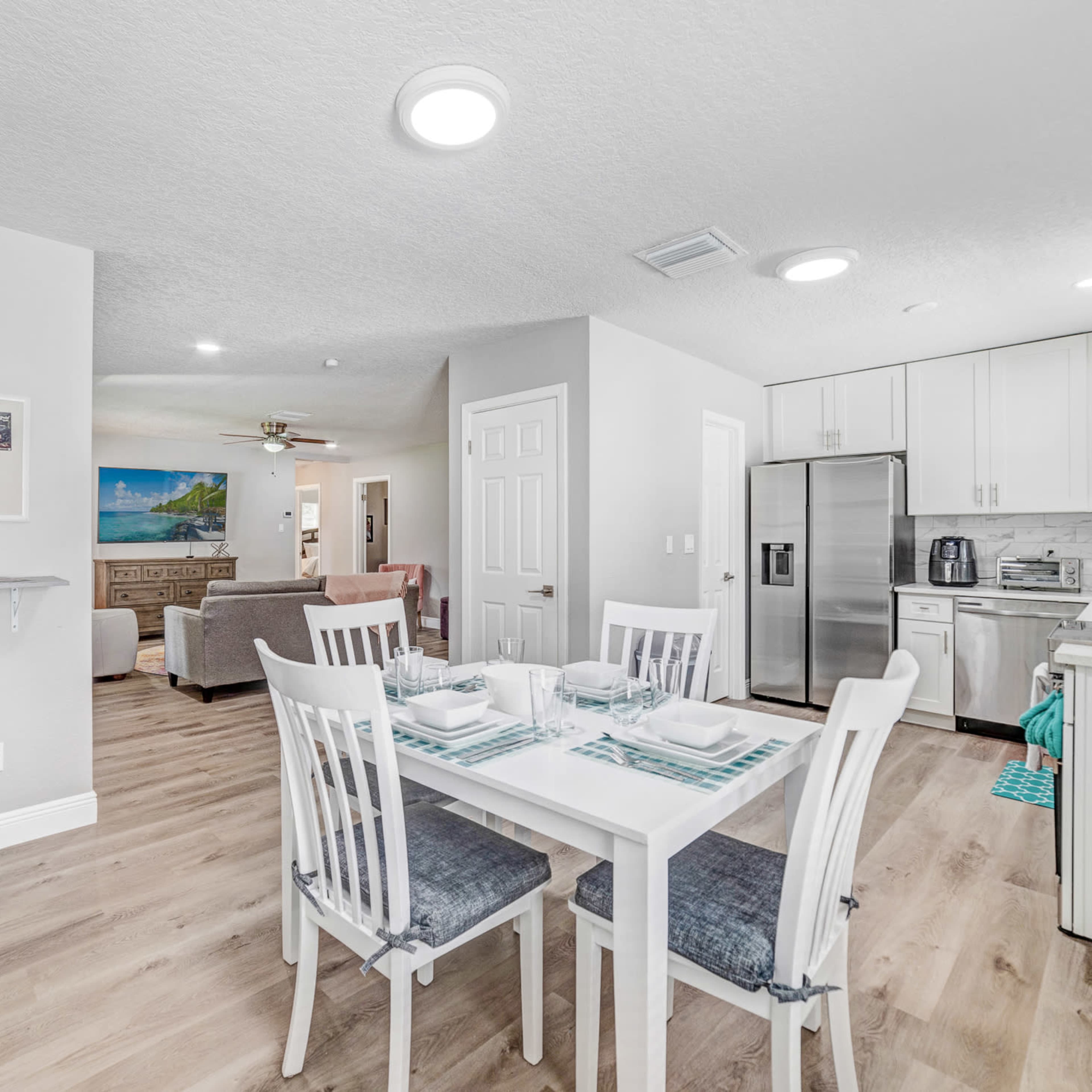 A modern, open-concept kitchen and dining area with a white table set for four, light-colored cabinetry, and a cozy living room visible in the background.