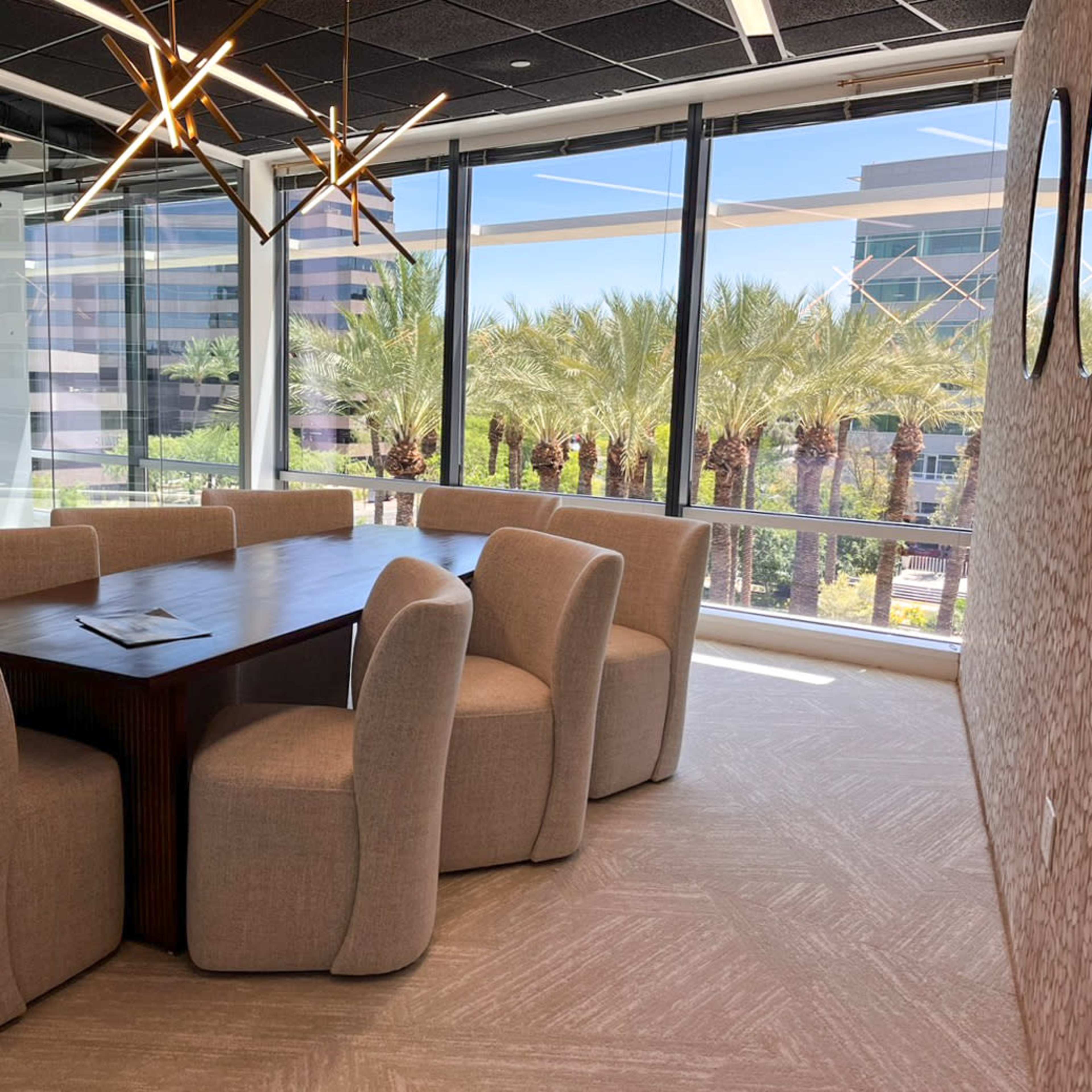 A modern conference room features a large wooden table surrounded by six upholstered chairs, with large windows offering a view of palm trees outside.