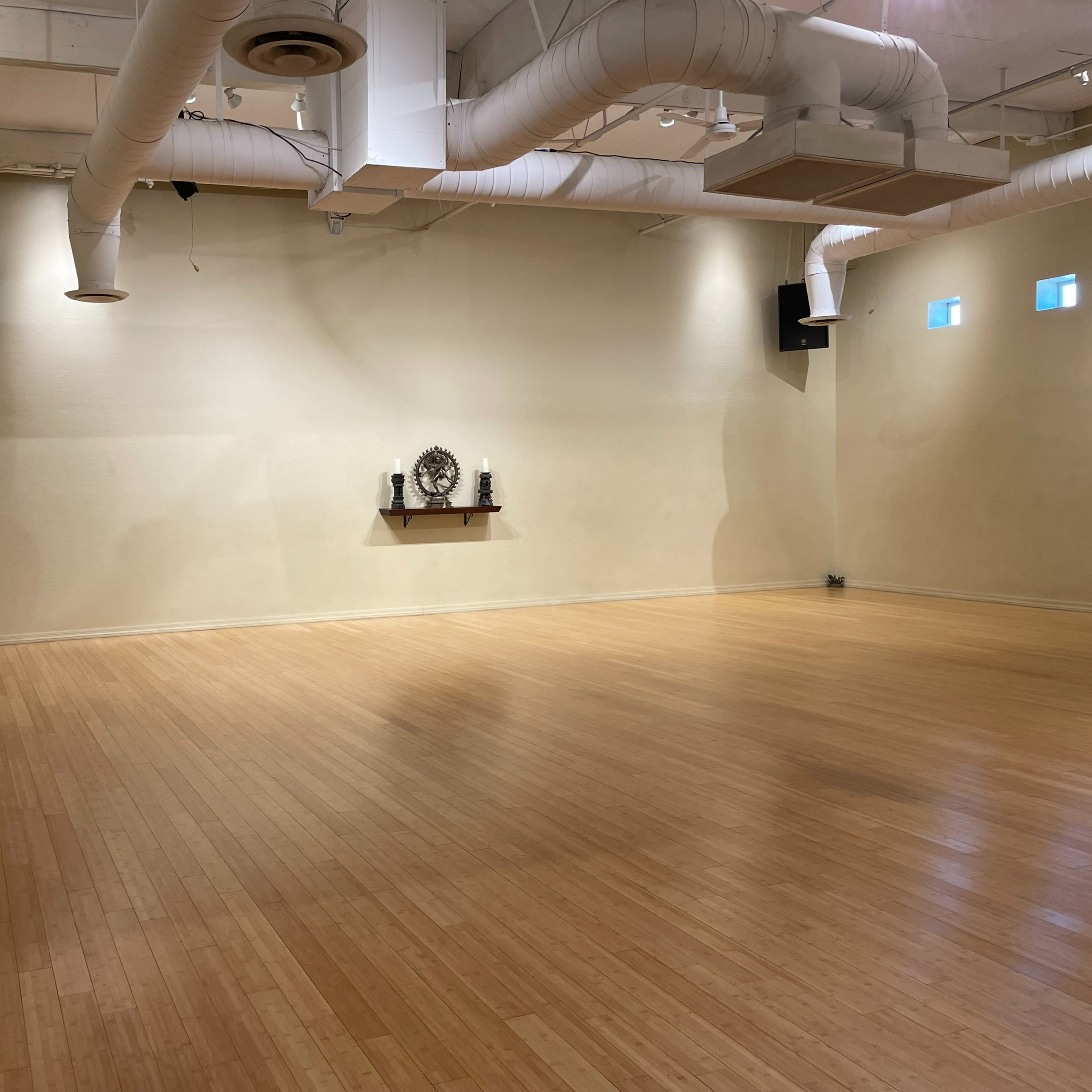 A spacious, empty studio with wooden flooring, beige walls, and a small shelf displaying decorative items.