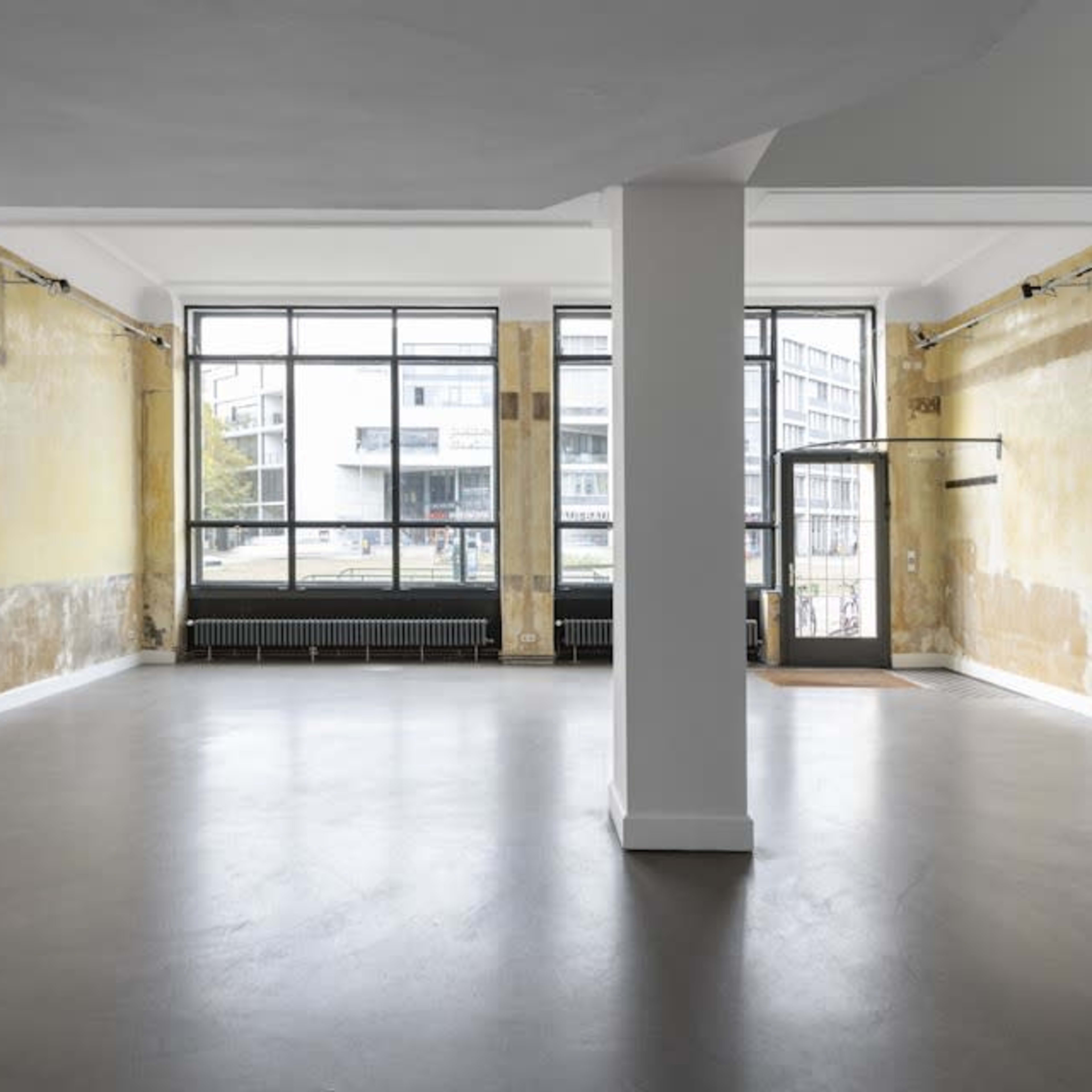 An empty room with large windows, featuring yellow-painted walls, a concrete floor, and a column in the center.