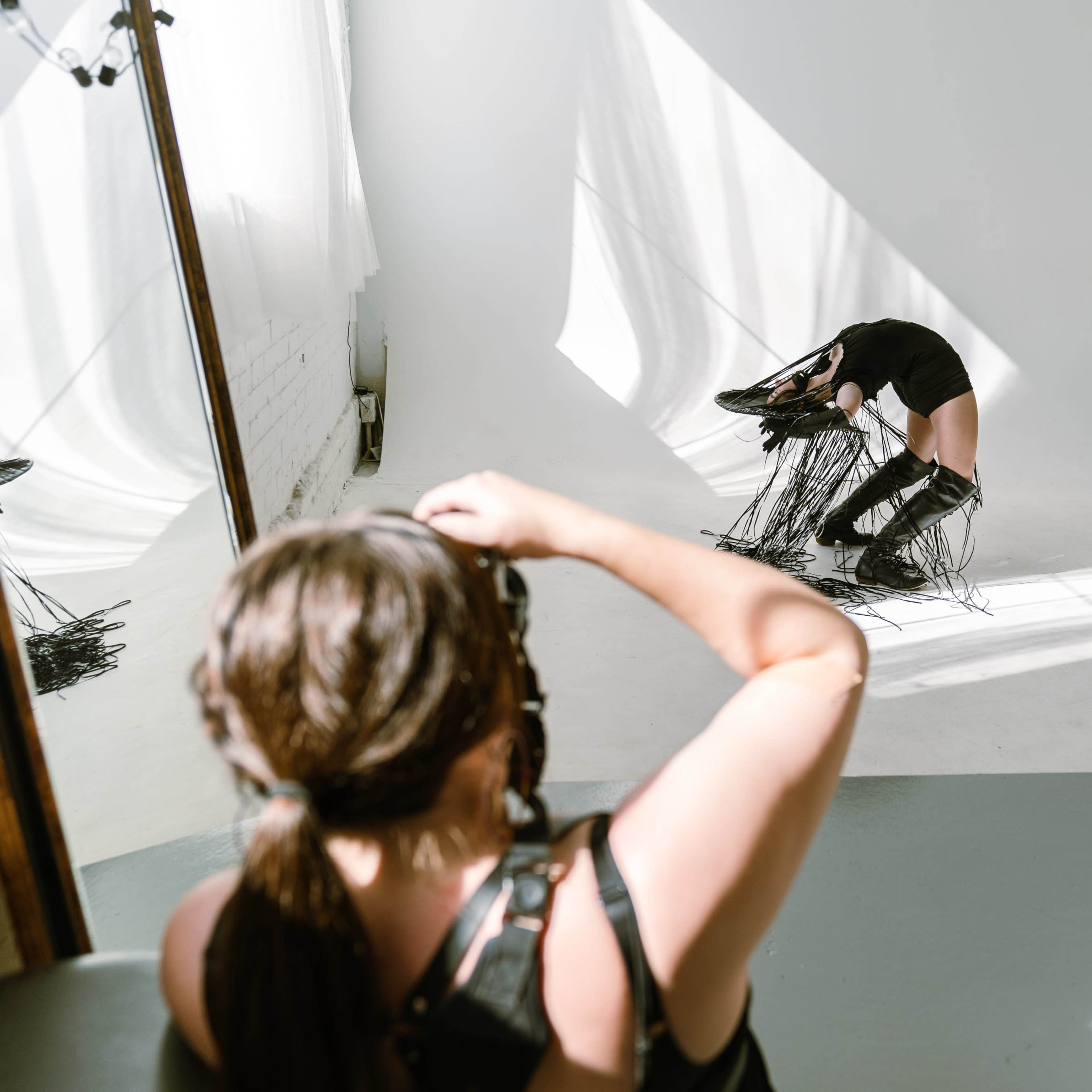 A model in a studio bending forward while wearing black clothing, with two photographers capturing the scene from different angles.