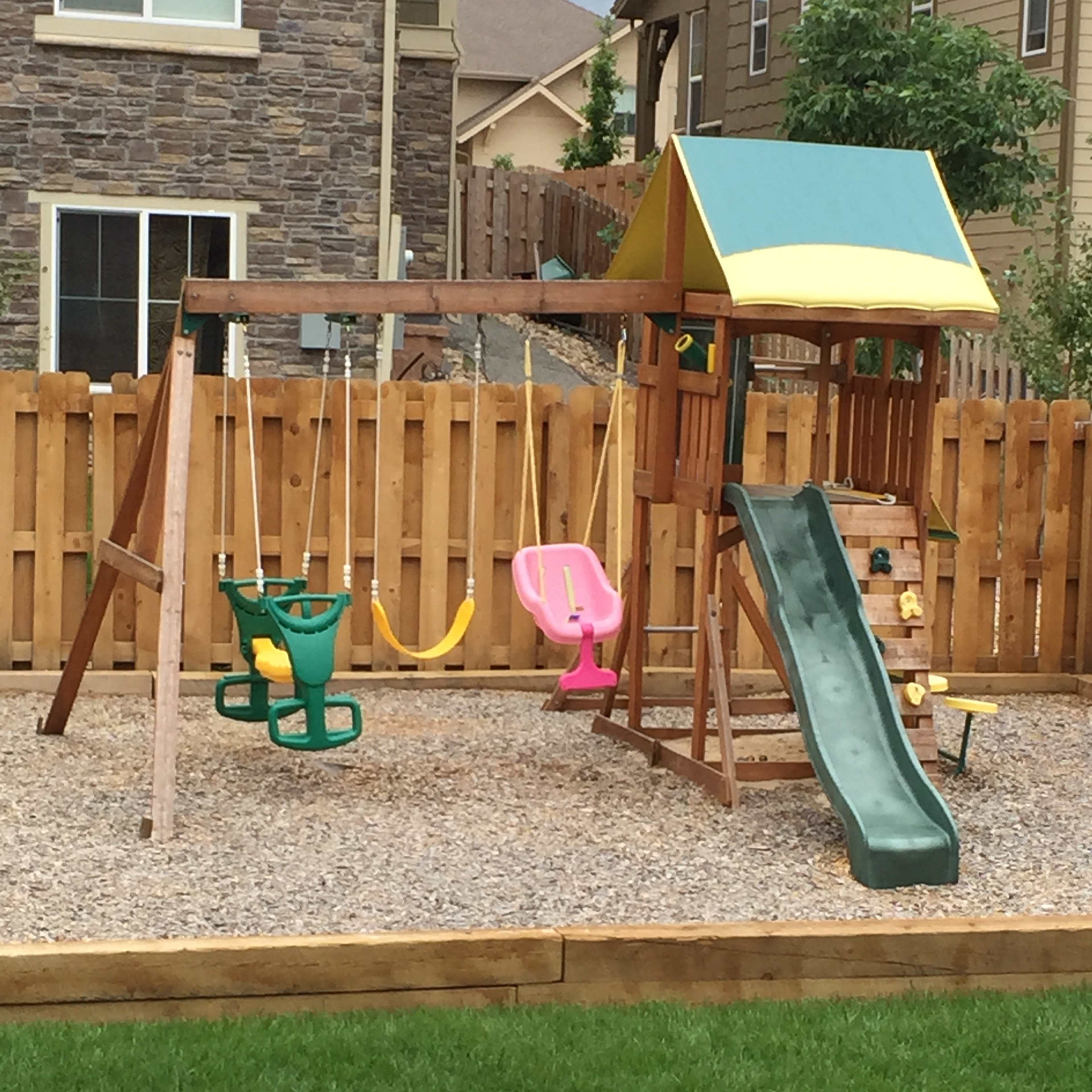 A wooden playground structure with a slide, swings, and a climbing area, surrounded by gravel and a wooden fence.