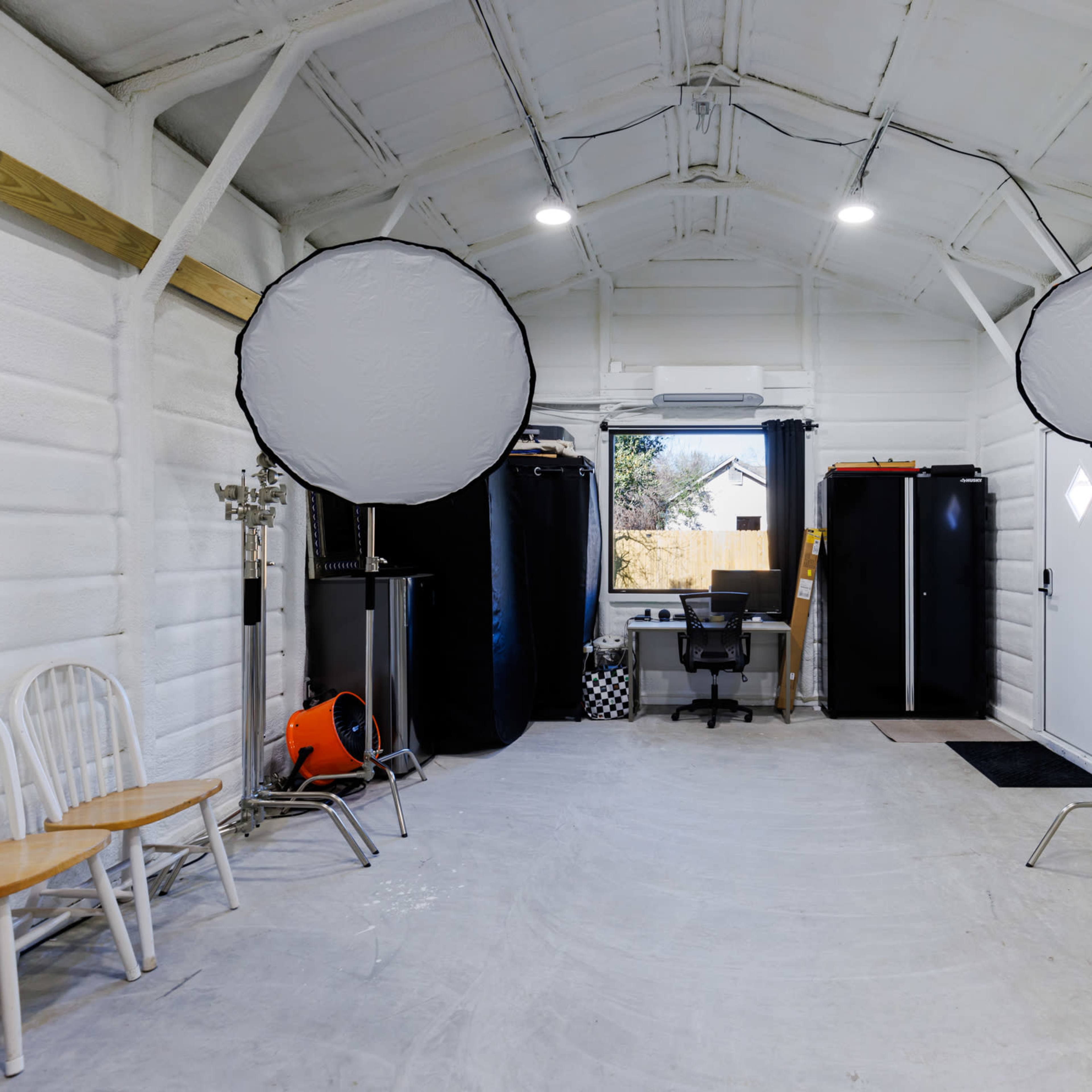 A spacious, well-lit interior of a garage or studio with two large softbox lights, a desk and chair, and two dark cabinets against the wall.